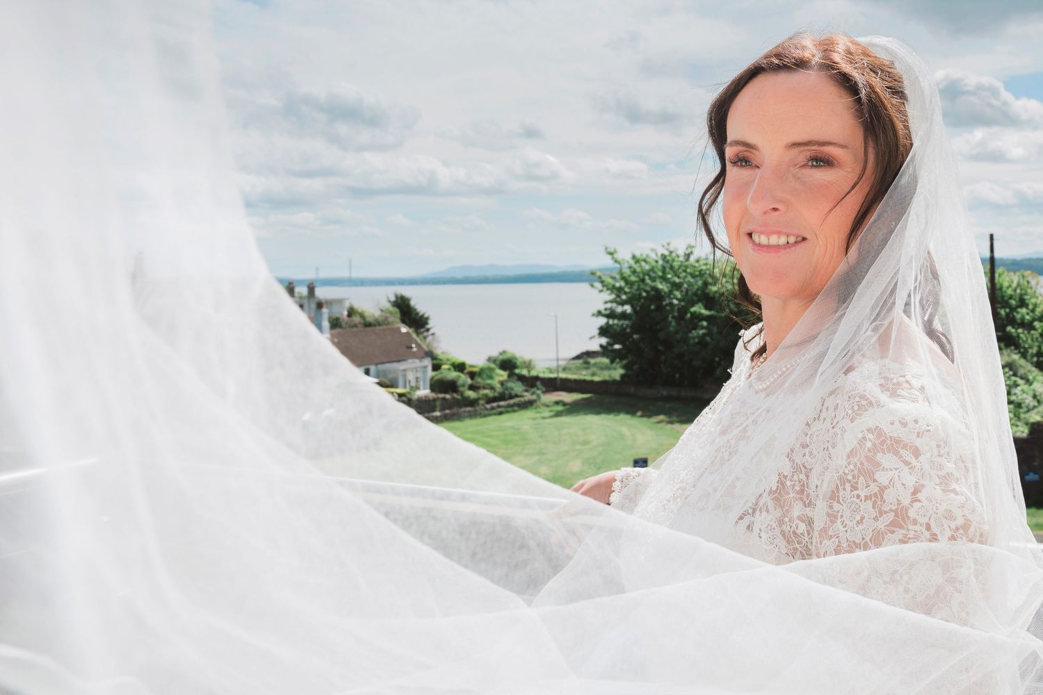Bride in lace wedding dress and flowing veil smiling outdoors with scenic water view and green garden background.