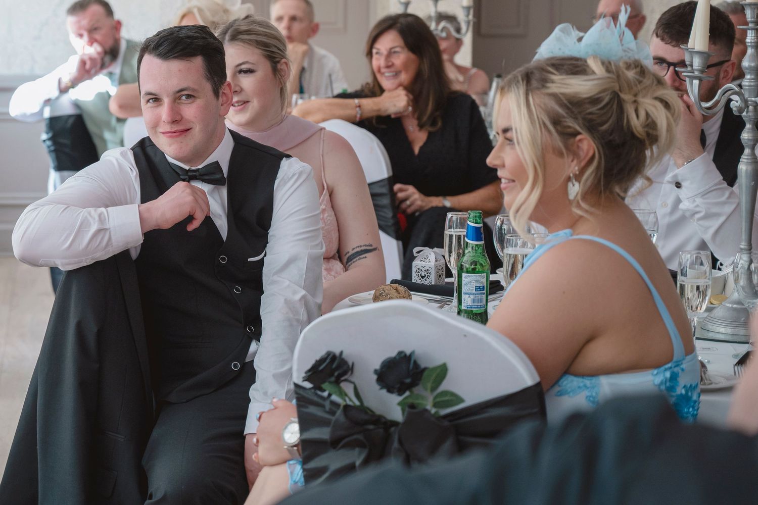 Wedding guests laughing and chatting at a reception table, bride in white gown beside groom in black tuxedo.