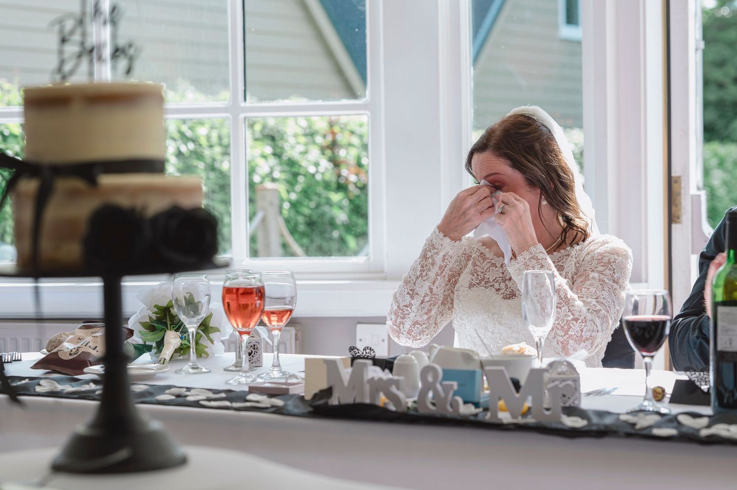 Emotional bride wiping tears at wedding reception table with cake, wine glasses, and decorative letters in bright venue.