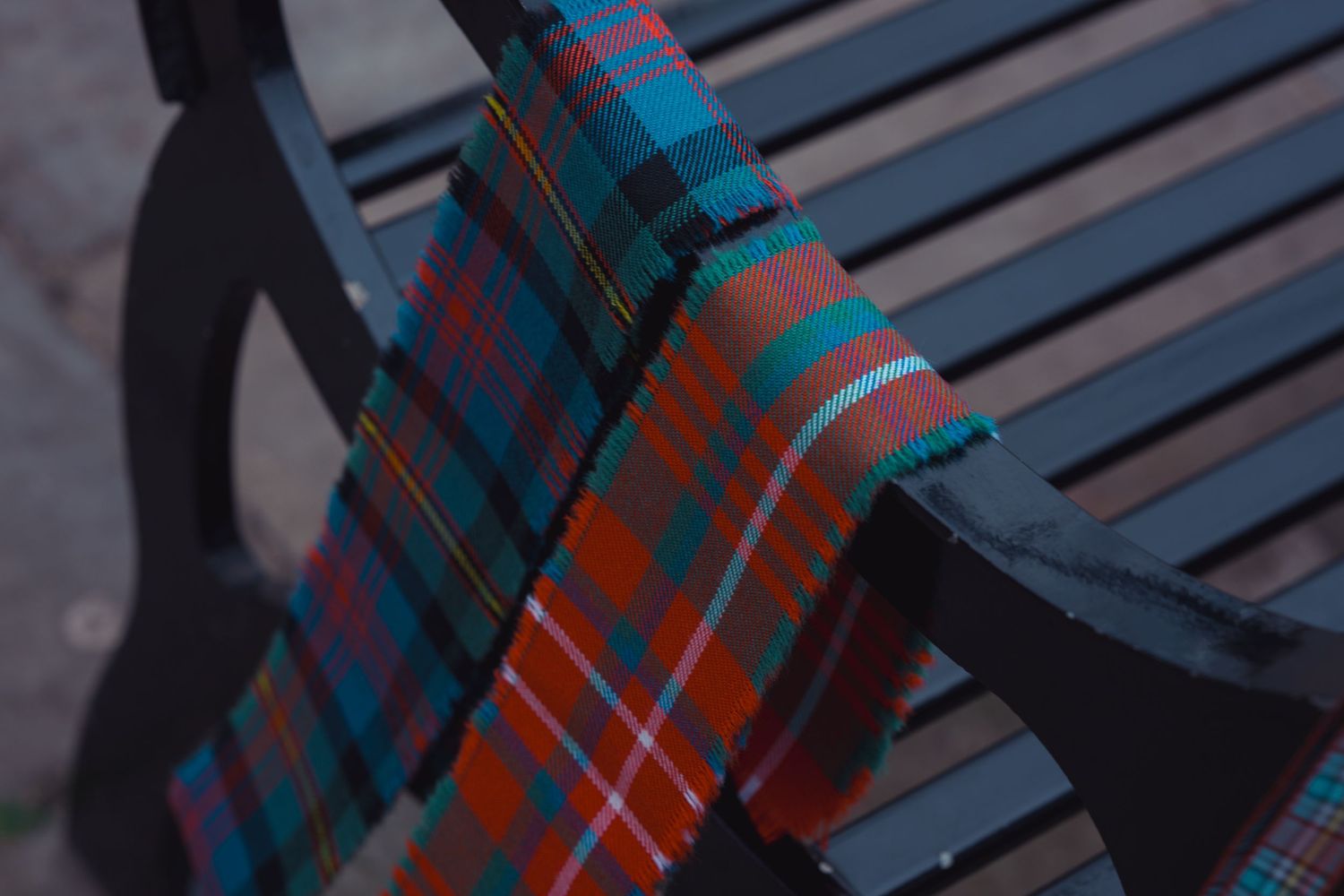 Red and teal tartan plaid scarf draped over a dark metal park bench outdoors.