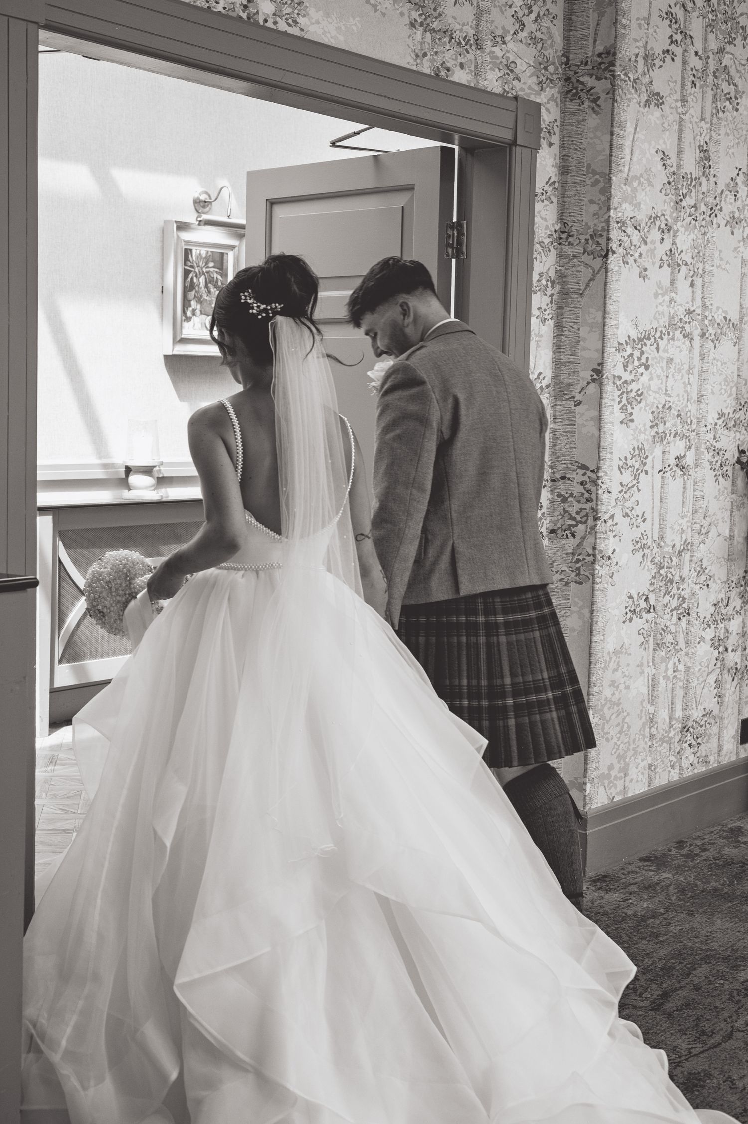 Bride in flowing white gown walks through elegant hallway with groom in Scottish attire.