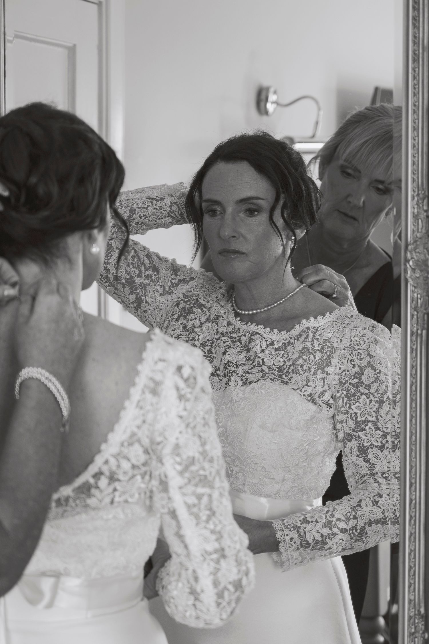 Black and white photo of a bride in lace gown being helped by a woman, reflected in an ornate mirror.