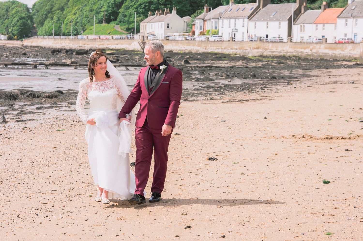 Bride and groom walking hand in hand on a sandy beach with a coastal village backdrop on their wedding day.