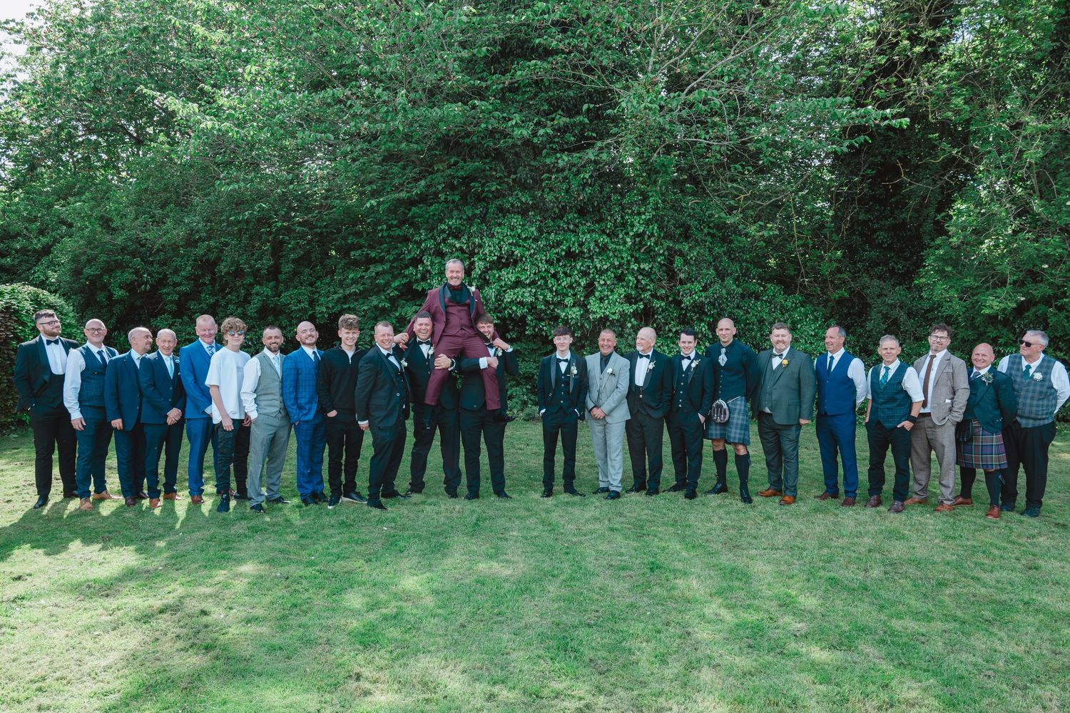 Large wedding party of groomsmen in blue suits posing outdoors on green lawn with lush trees in background.