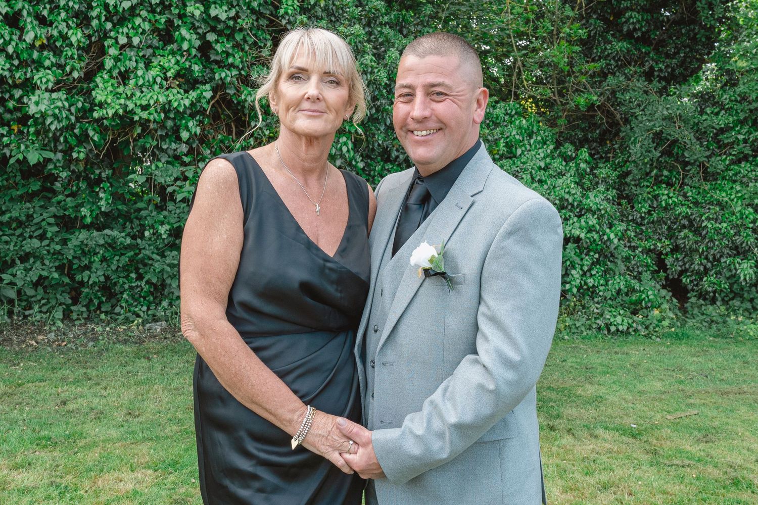Elegant couple posing outdoors, woman in black dress and man in gray suit with boutonniere, smiling at a formal event.