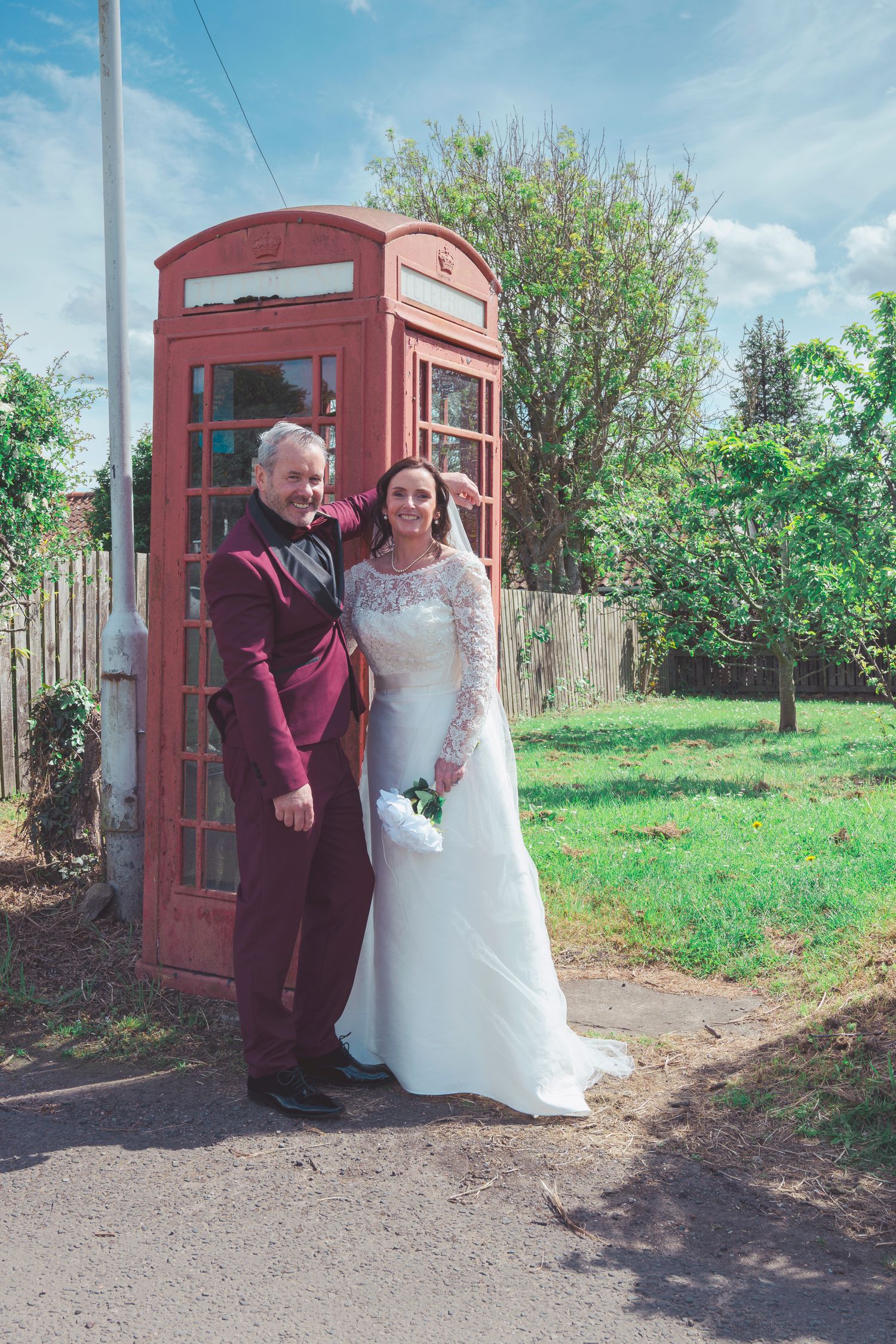 Bride and groom posing together in front of a red British phone box on a sunny day.