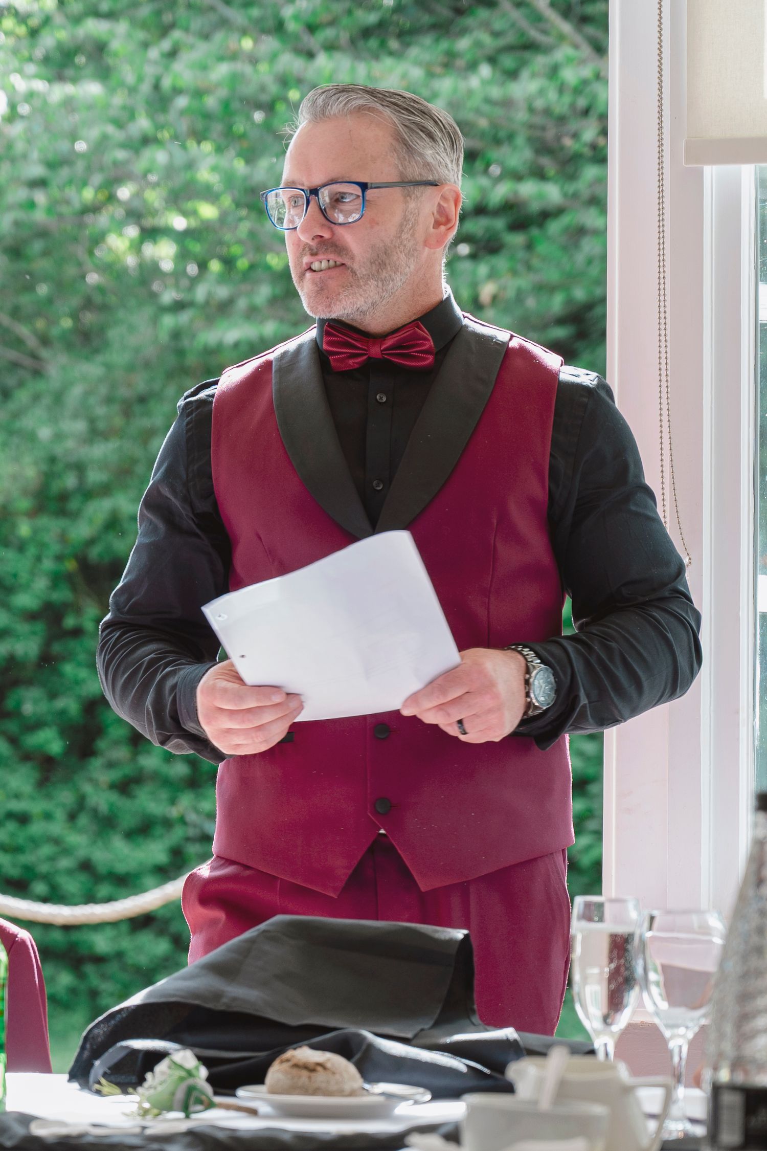 Elegant middle-aged man in red vest and bow tie holds papers while speaking at a formal dining event.