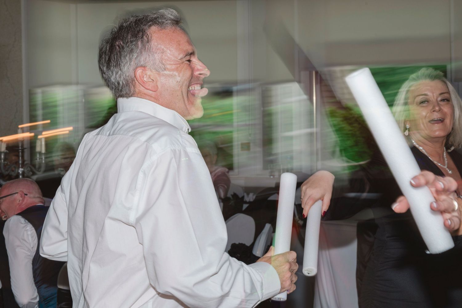 Man in white shirt laughing while holding white batons at an indoor event with guests in background.