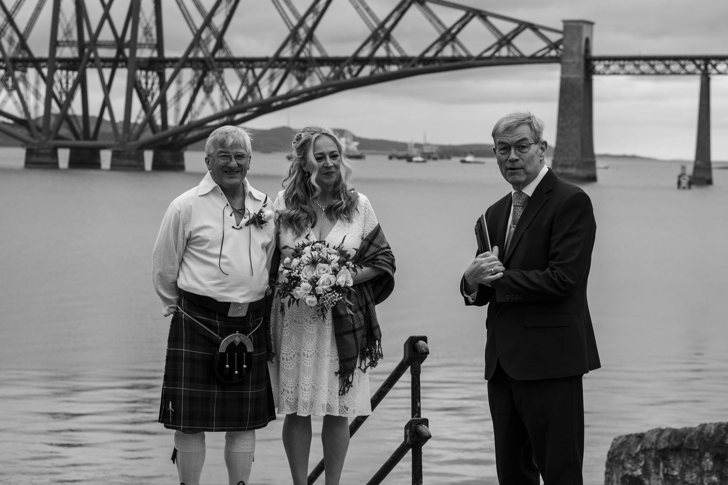 Black and white wedding photo of bride and two men in kilts by the Forth Rail Bridge in Scotland.