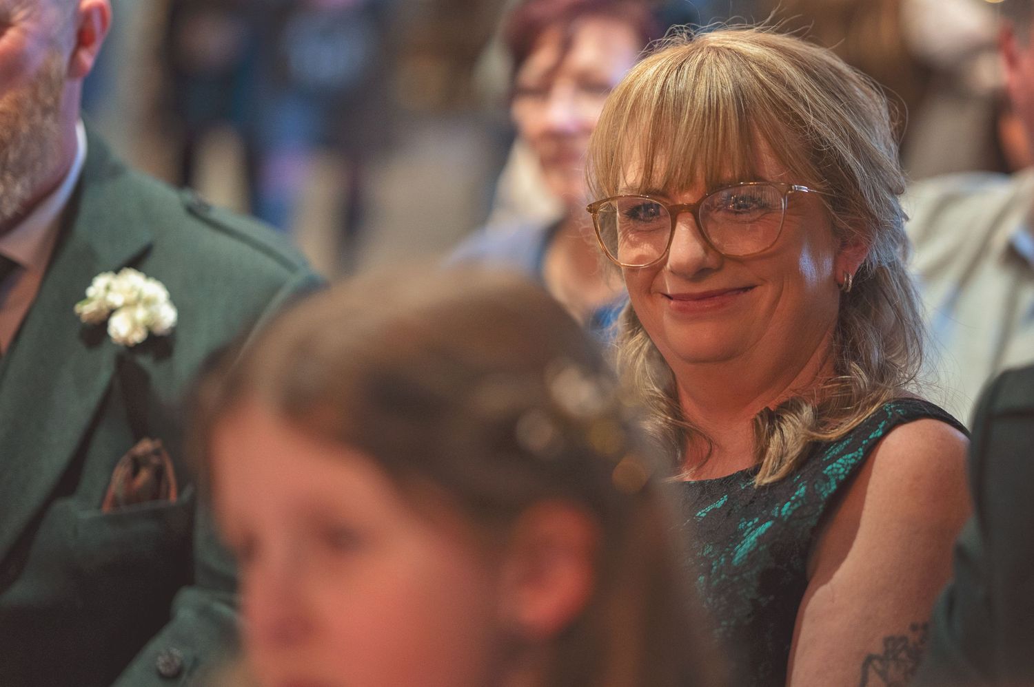 Smiling woman with glasses wearing teal dress watches emotional wedding ceremony from guest seating area.