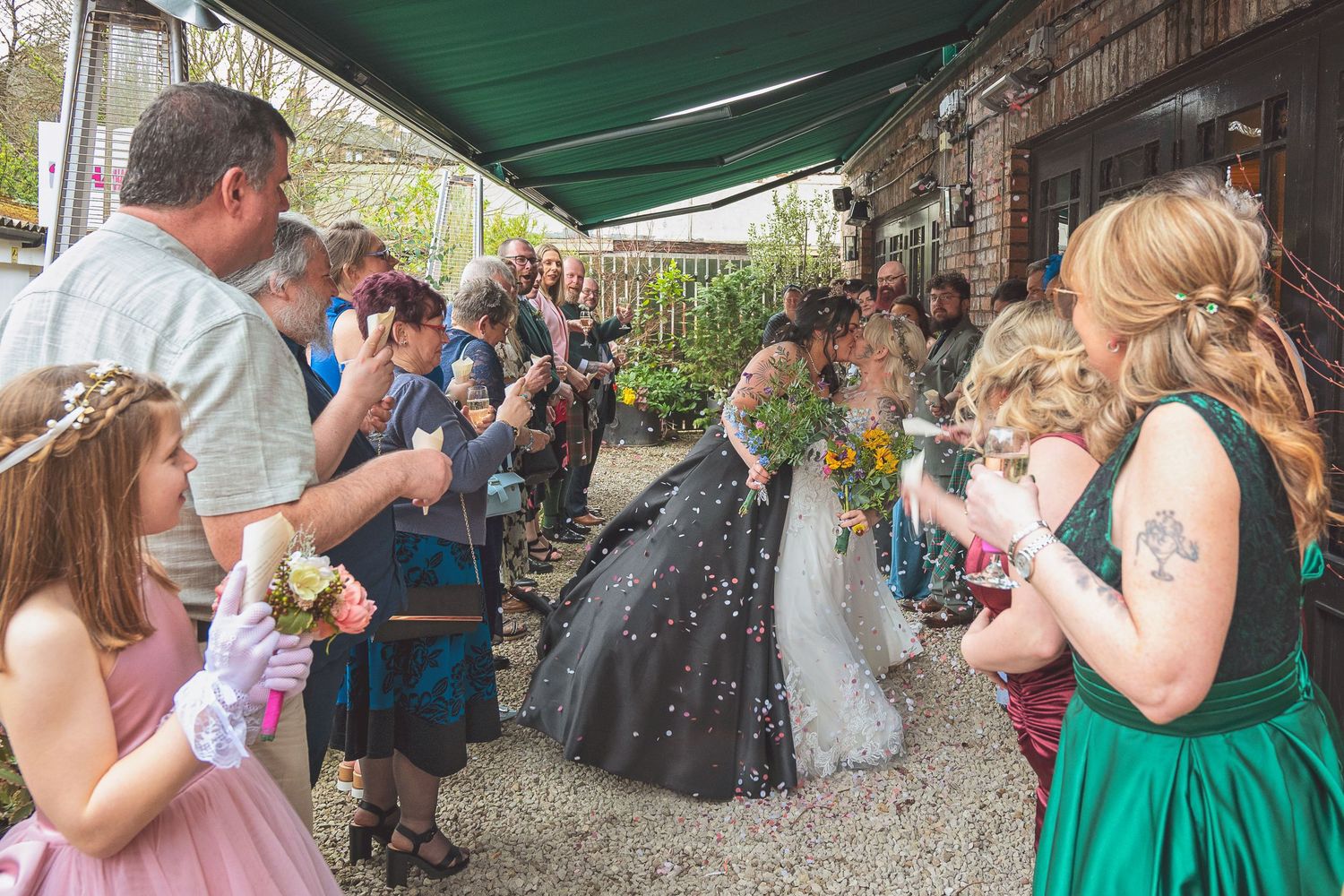 Wedding guests blowing bubbles as two brides in black and white gowns exit ceremony.