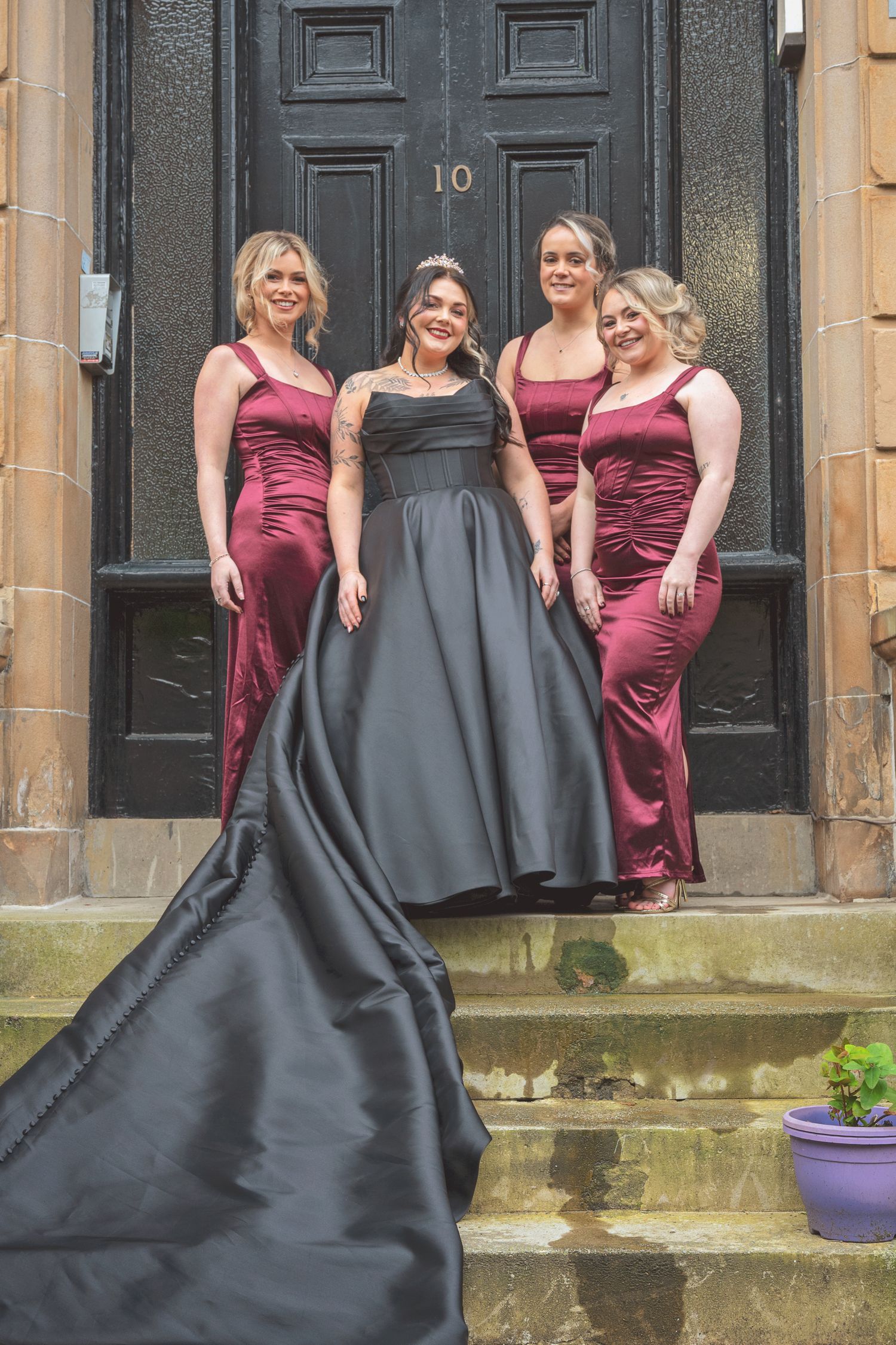 Bride in black ball gown with train poses with three bridesmaids in burgundy satin dresses on stone steps.