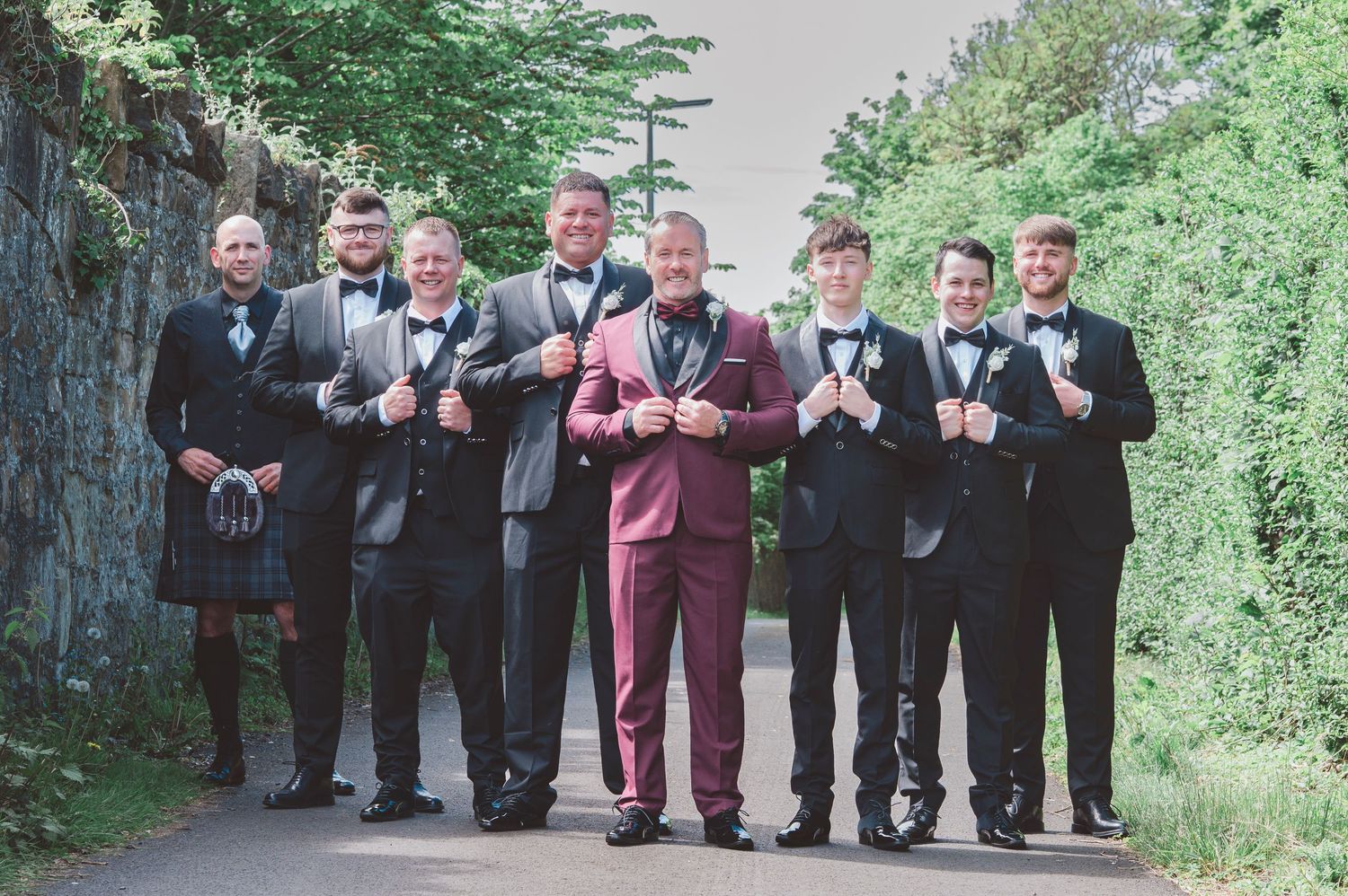 Groom in burgundy suit stands with groomsmen in black tuxedos and one in a kilt on a scenic outdoor path.