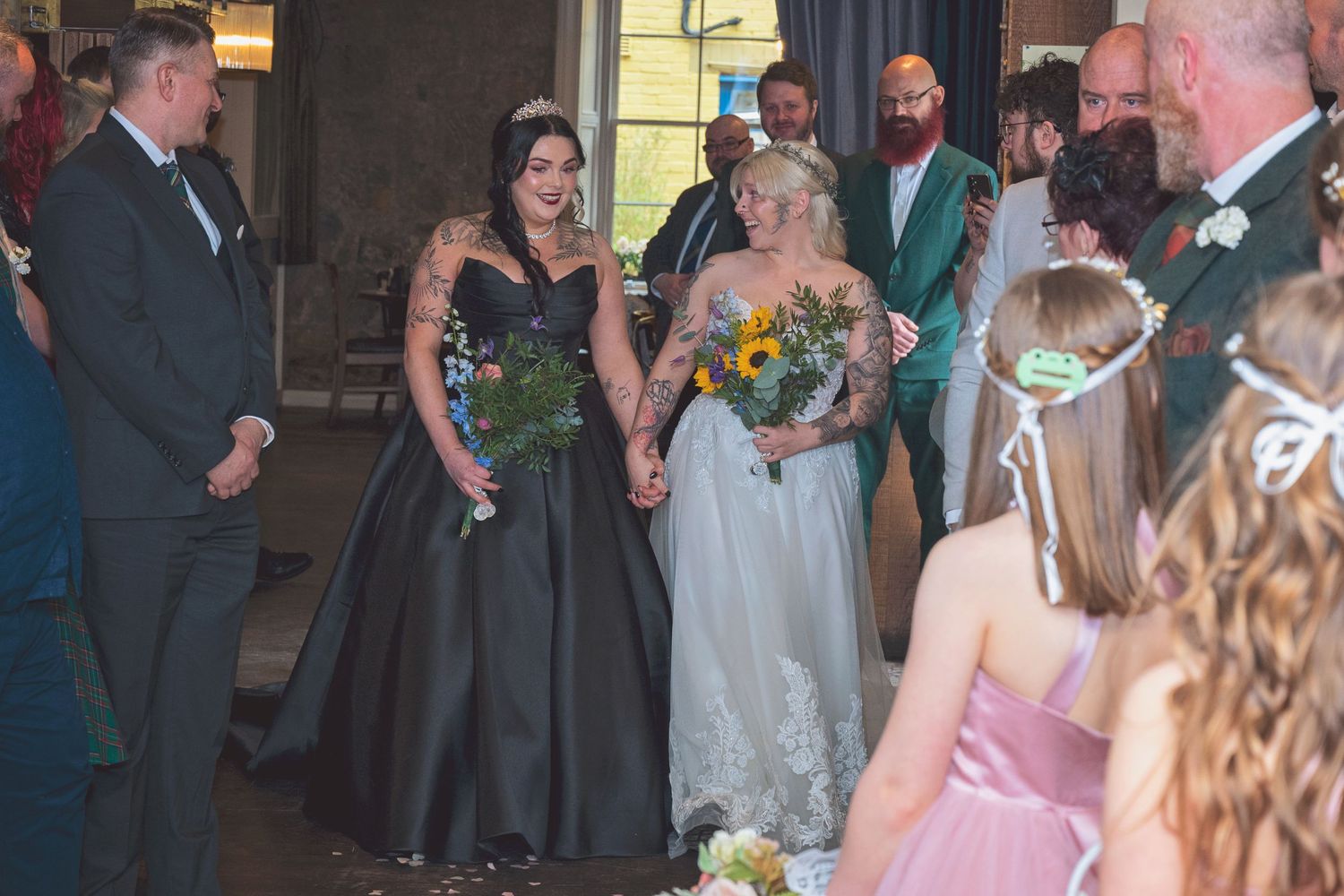 Two brides walk down aisle holding bouquets in black and white gowns at wedding ceremony.