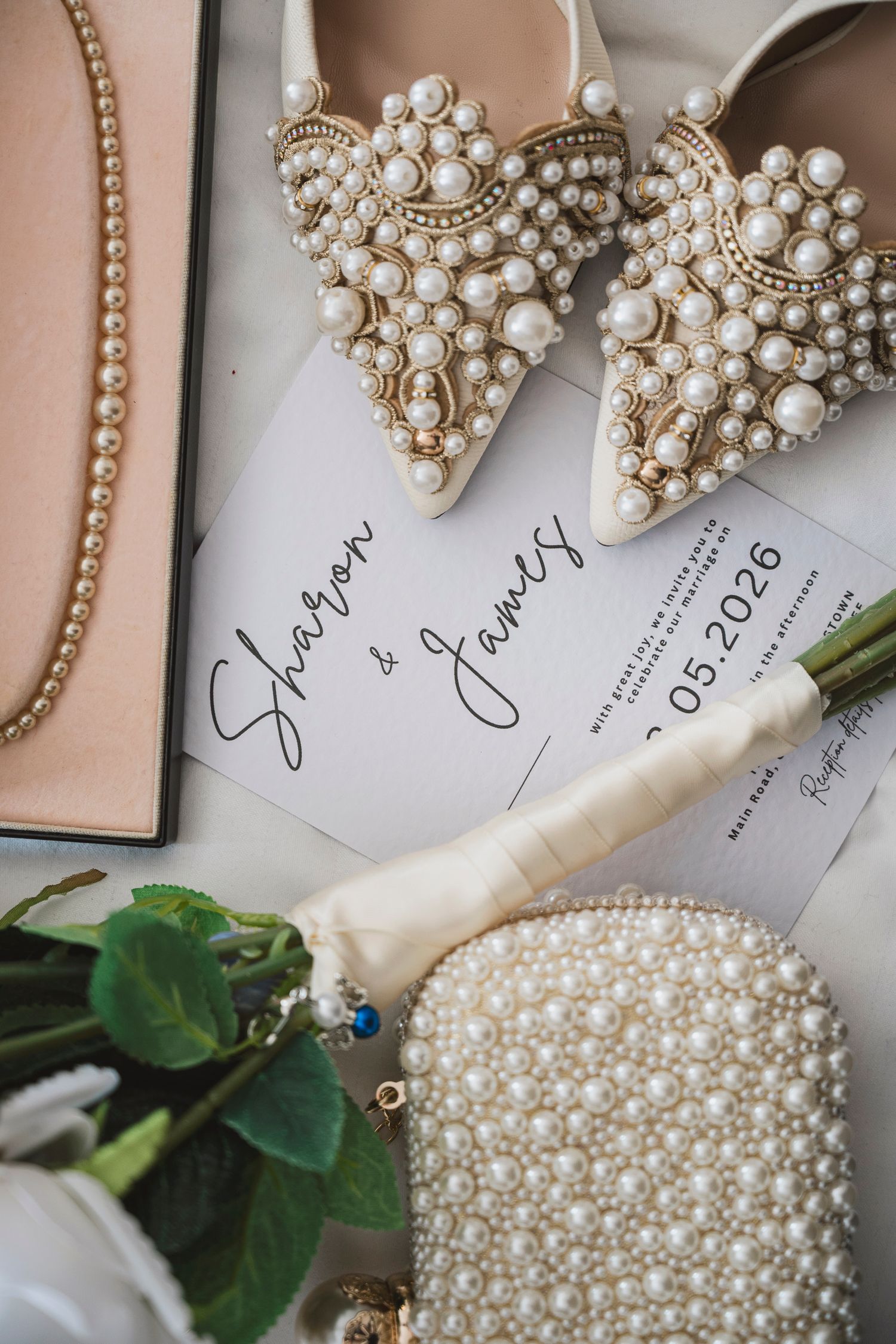 Pearl-embellished bridal heels and clutch beside Sharon and James wedding invitation with white roses flatlay.