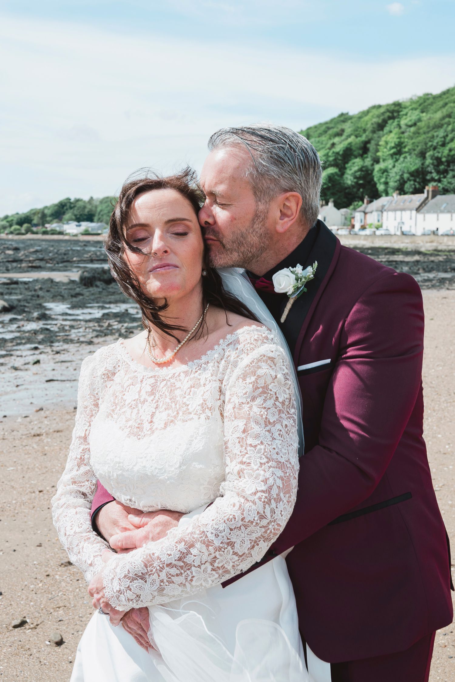 Groom in burgundy suit kissing bride in lace wedding dress on a sandy beach with green trees behind.