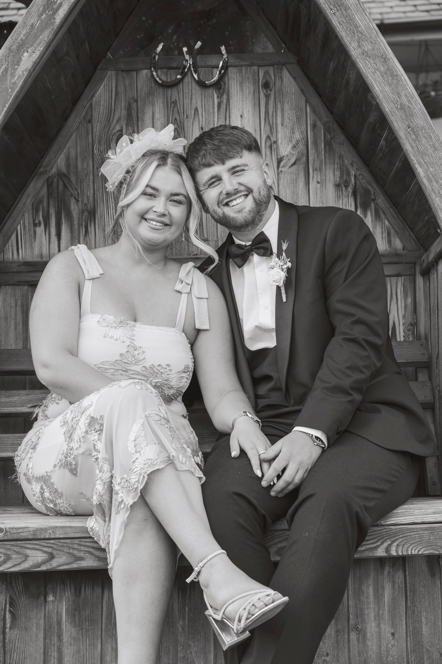 Black and white photo of a smiling couple in wedding attire sitting together in a wooden gazebo.