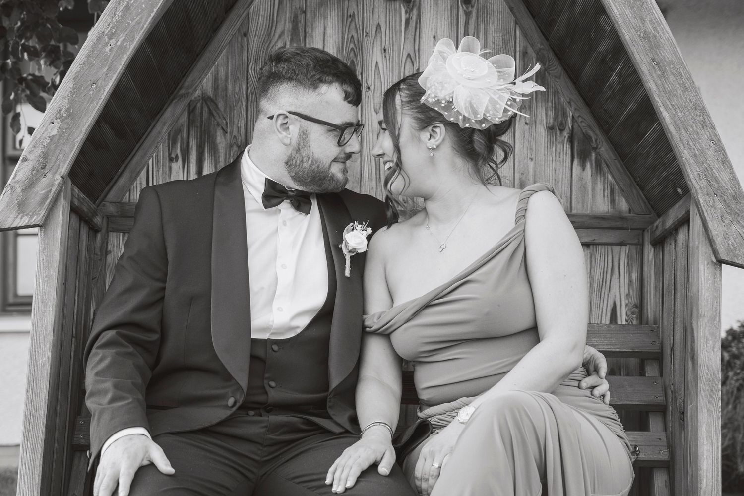 Black and white photo of a couple in wedding attire sitting together under a wooden arch, sharing a romantic moment.