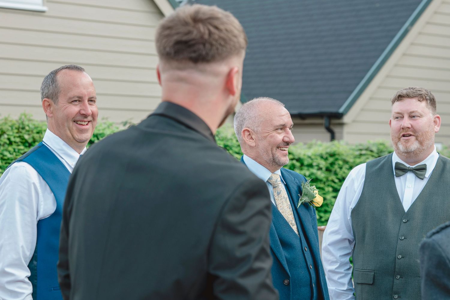 Groomsmen in blue waistcoats and bow ties laughing together outside a wedding venue on a sunny day.