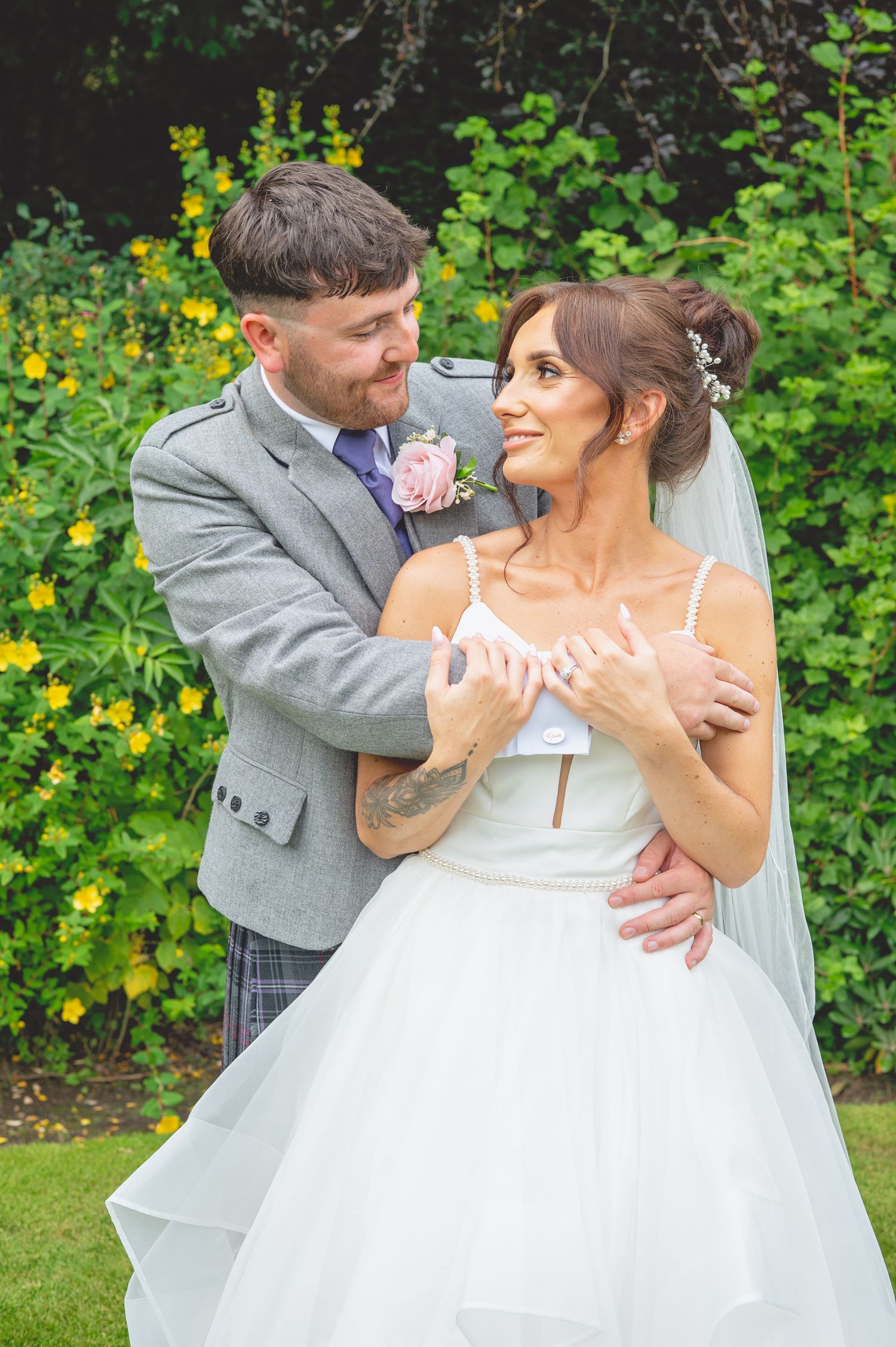Newlyweds embrace in a garden with yellow flowers and greenery in the background.