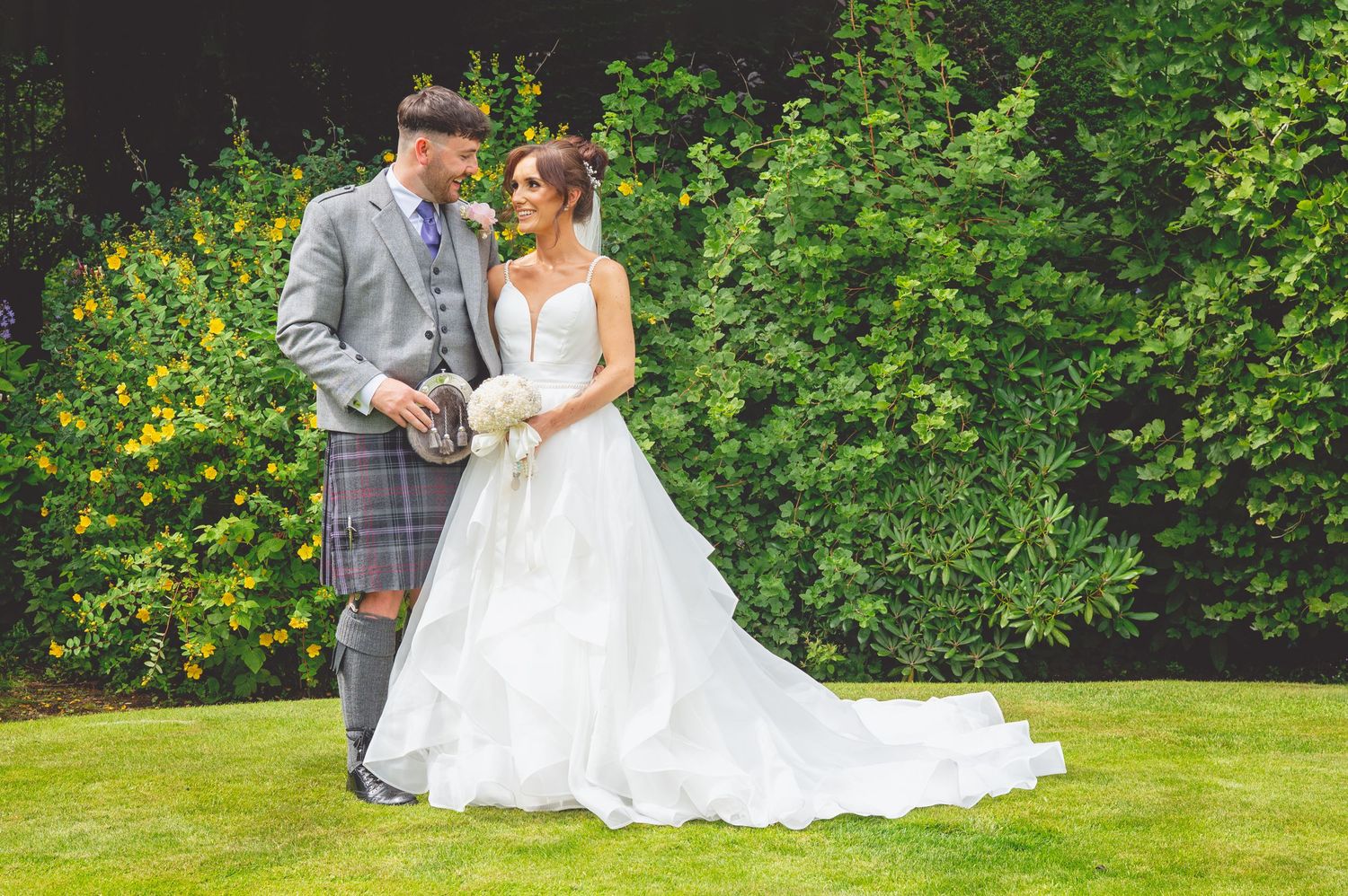 Wedding couple poses on manicured lawn with lush green garden backdrop.