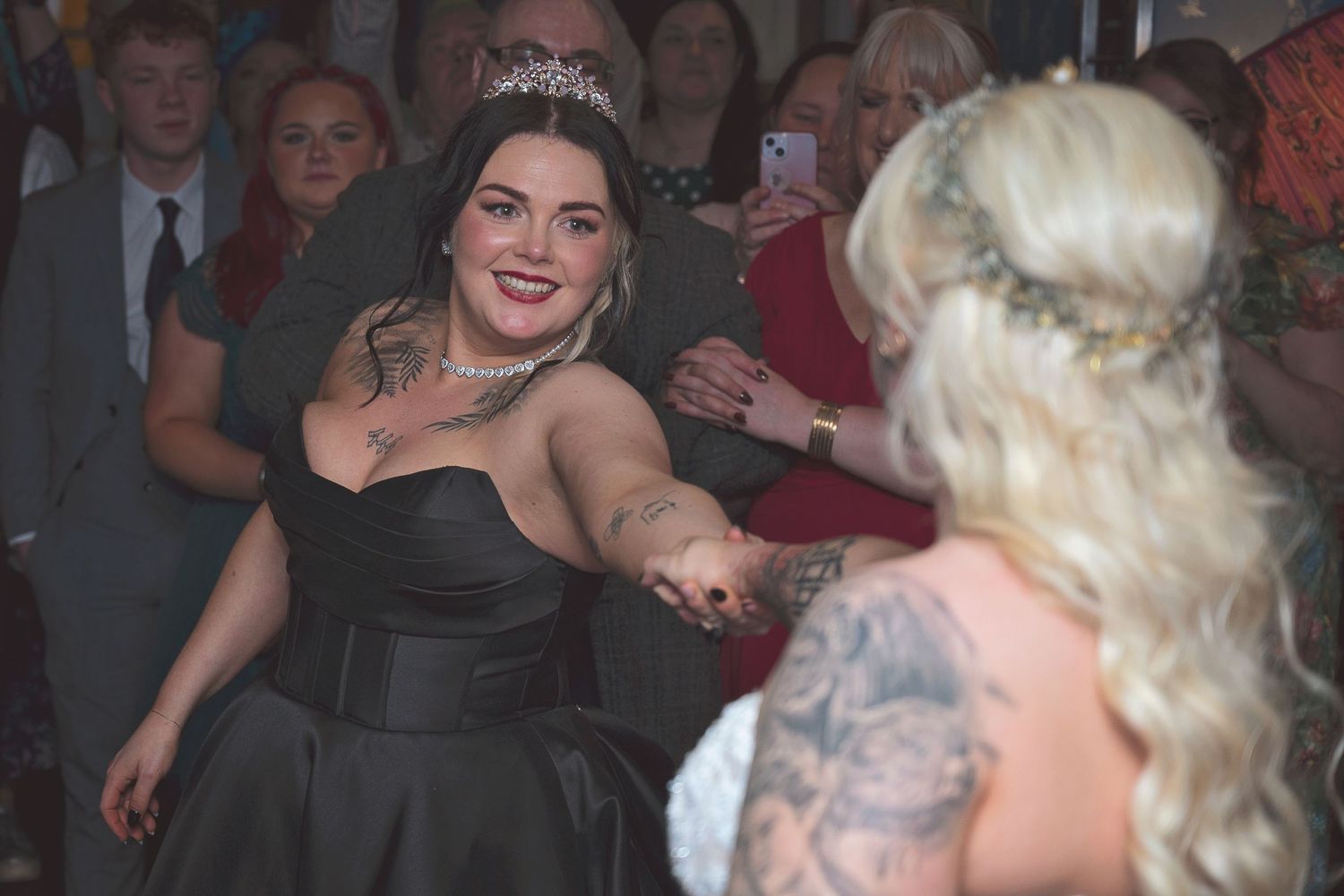 Two women in formal attire hold hands on a dance floor, one in black gown with tiara, other in white bridal dress.