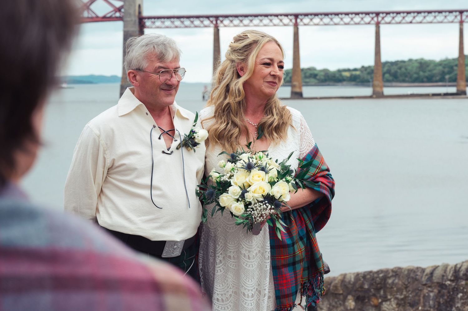 Bride holding white bouquet stands with older man near waterfront bridge at outdoor wedding ceremony.
