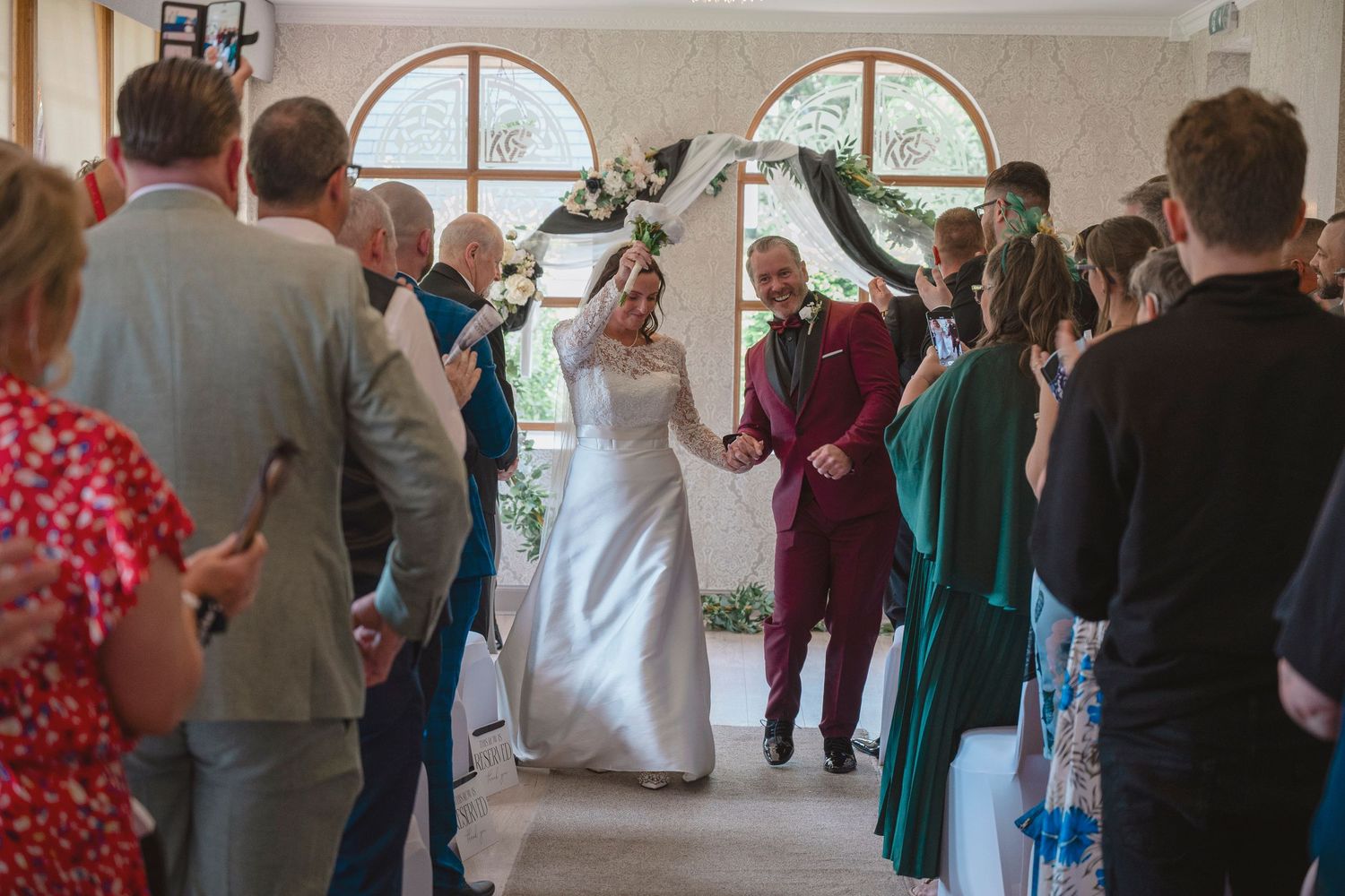 Bride and groom walk down the aisle together at an elegant indoor wedding ceremony with guests applauding.