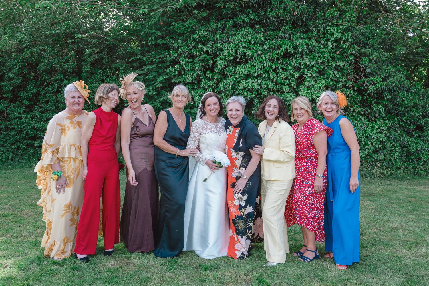A bride in white poses with eight colorful guests outdoors at a wedding celebration surrounded by lush green trees.