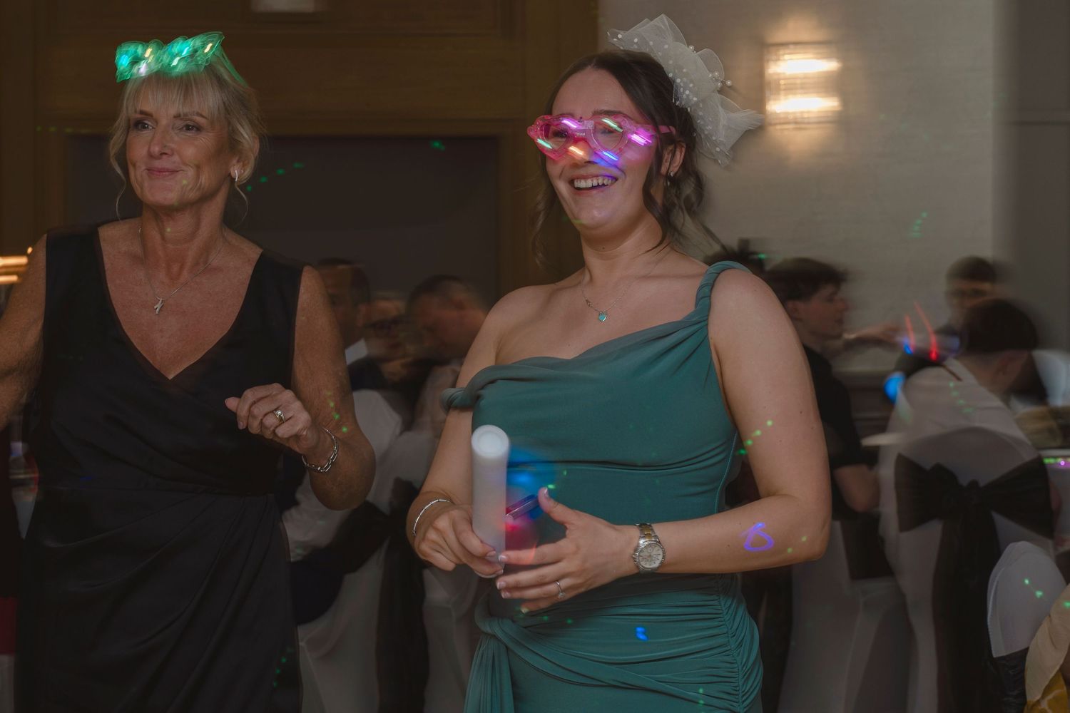 Two women dancing at a formal event, one wearing pink star-shaped glasses and holding a glow stick.