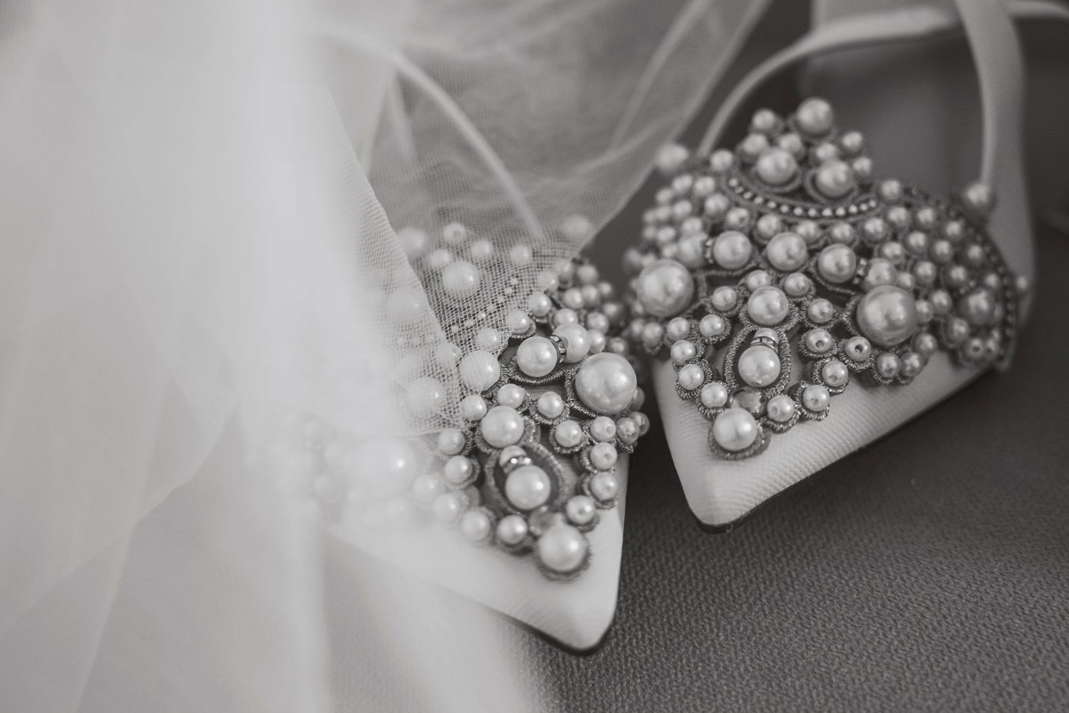 Black and white photo of pearl-embellished bridal heels beside a wedding veil on a gray surface.