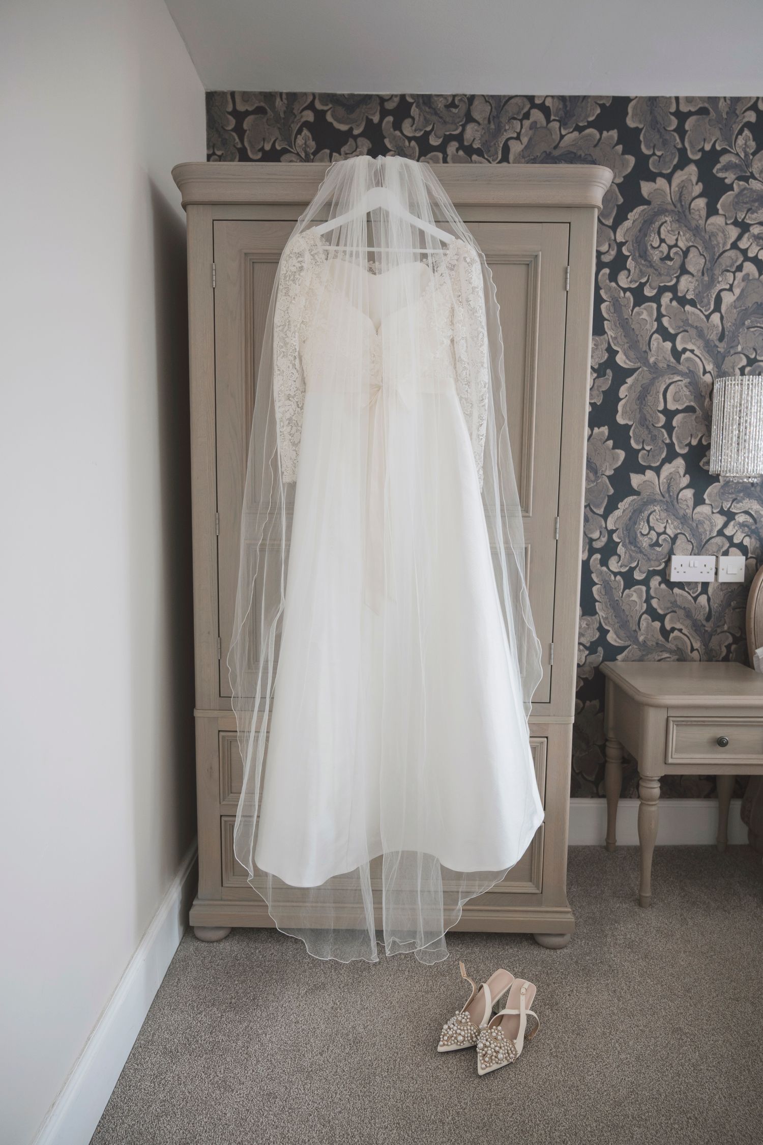 White lace wedding dress with veil hanging on wardrobe, bridal heels on floor below.