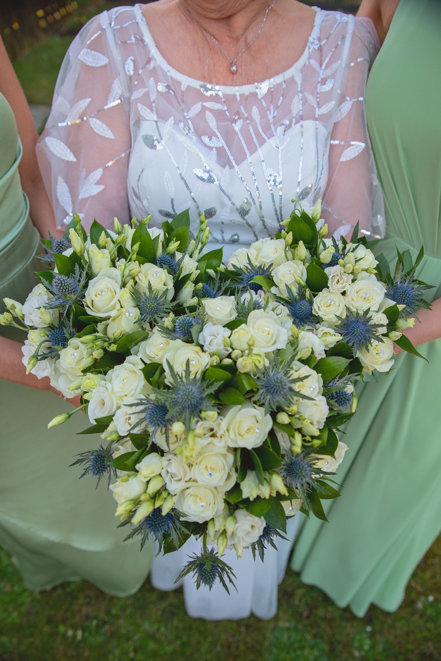 A wedding bouquet featuring white roses and blue thistles held against a mint green dress.