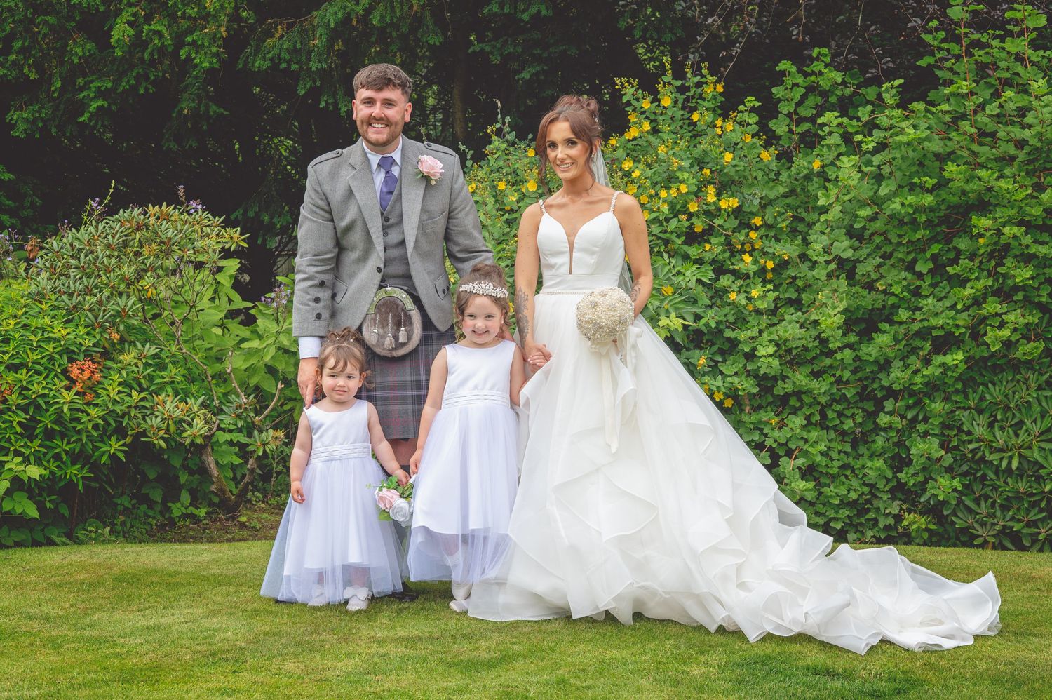 Wedding portrait with couple and two children in matching formal attire on garden lawn.
