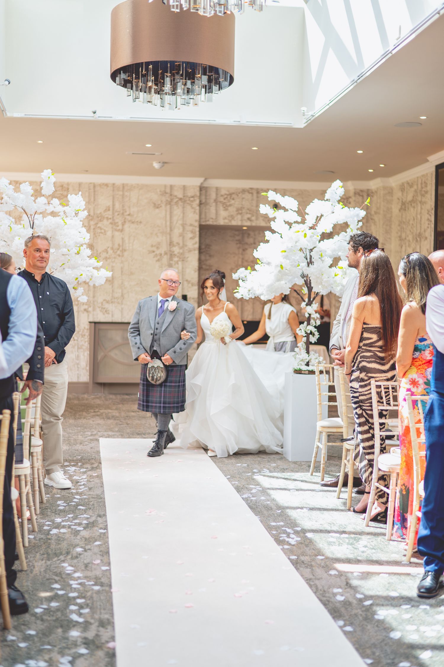 Wedding ceremony in an elegant venue with Scottish kilts and white floral decorations along the aisle.