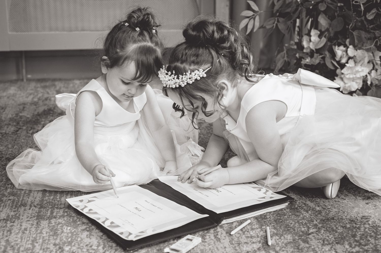 Young attendants in white dresses sign a book together on the floor.