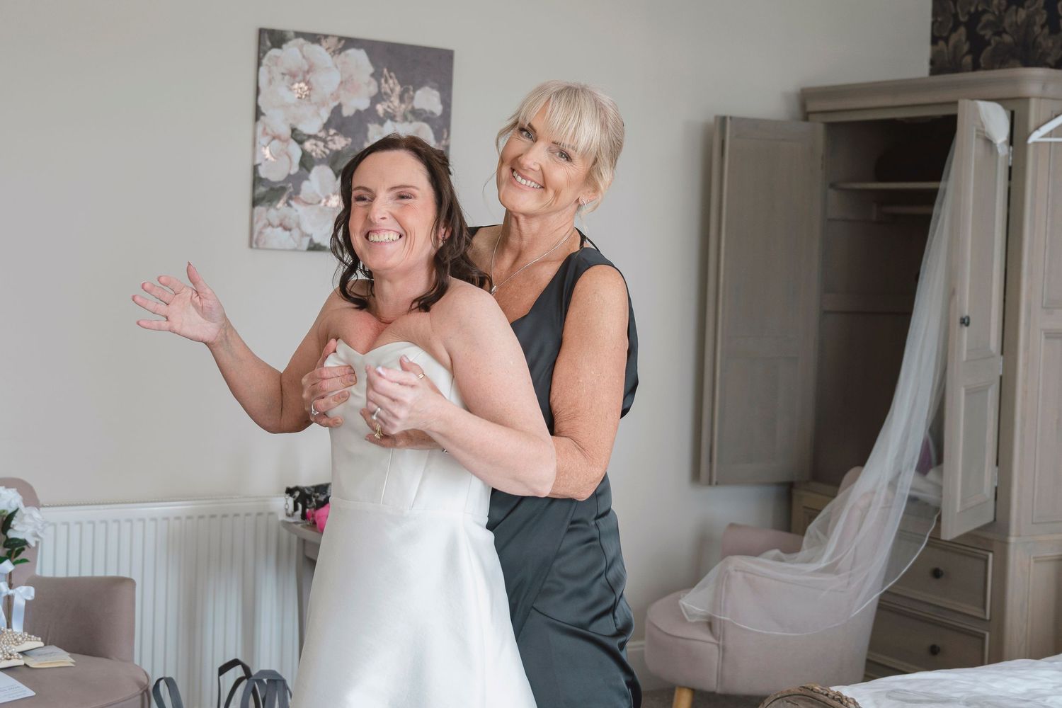 Bride in white dress being helped by smiling blonde woman in grey dress in a bright wedding preparation room.