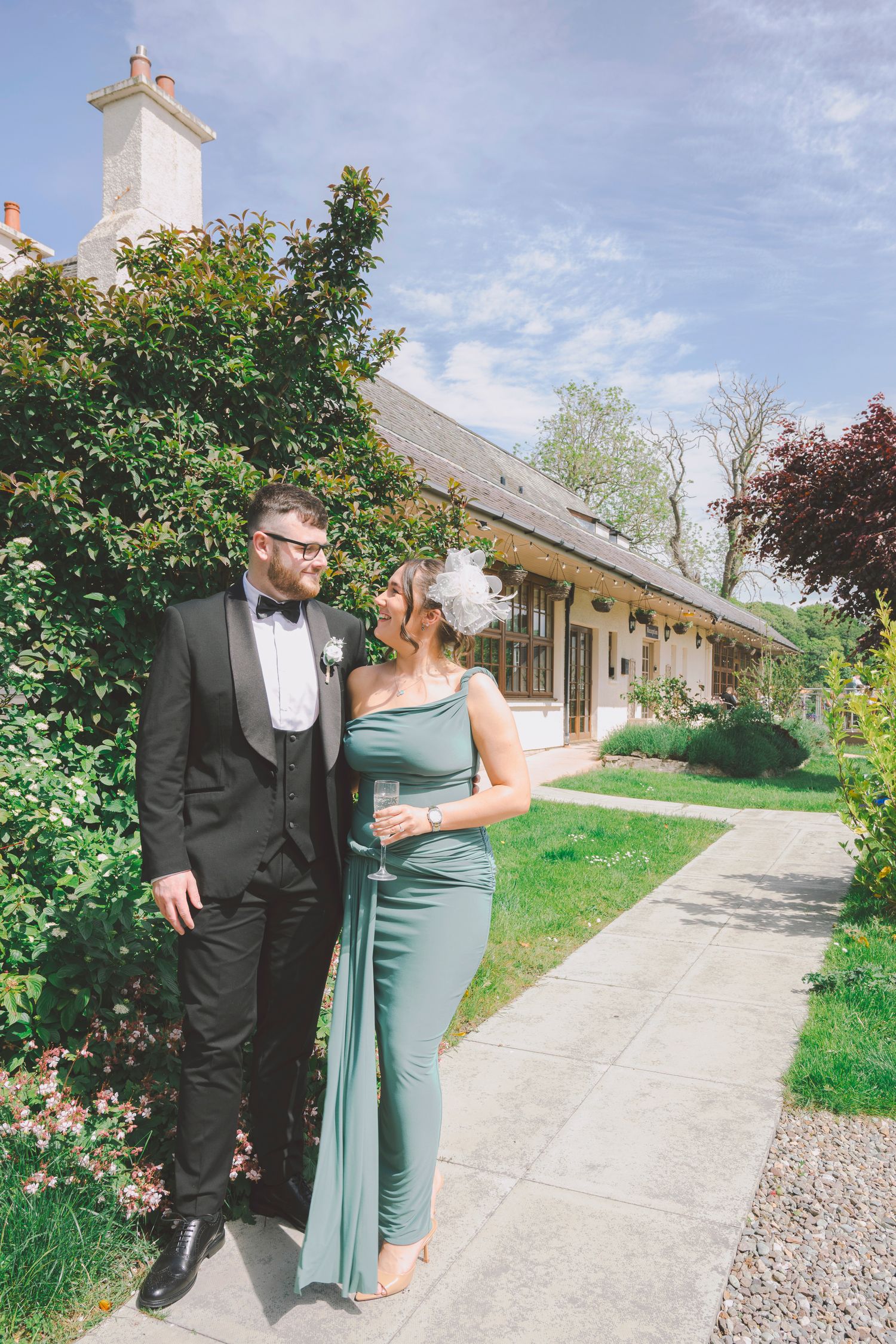 Couple in formal attire, man in black tuxedo and woman in sage green gown, posing outdoors at a wedding venue.