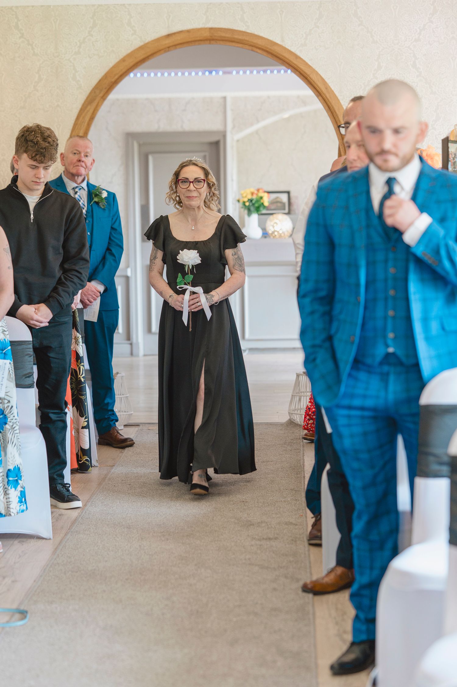 Woman in black dress walks down wedding aisle holding white flower, guests and groom in blue suit visible.