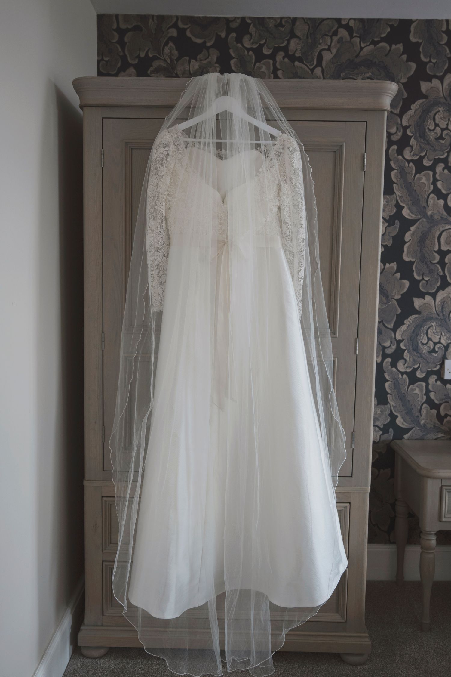 Elegant white lace wedding dress with long veil hanging on a wooden armoire in a decorated bedroom.