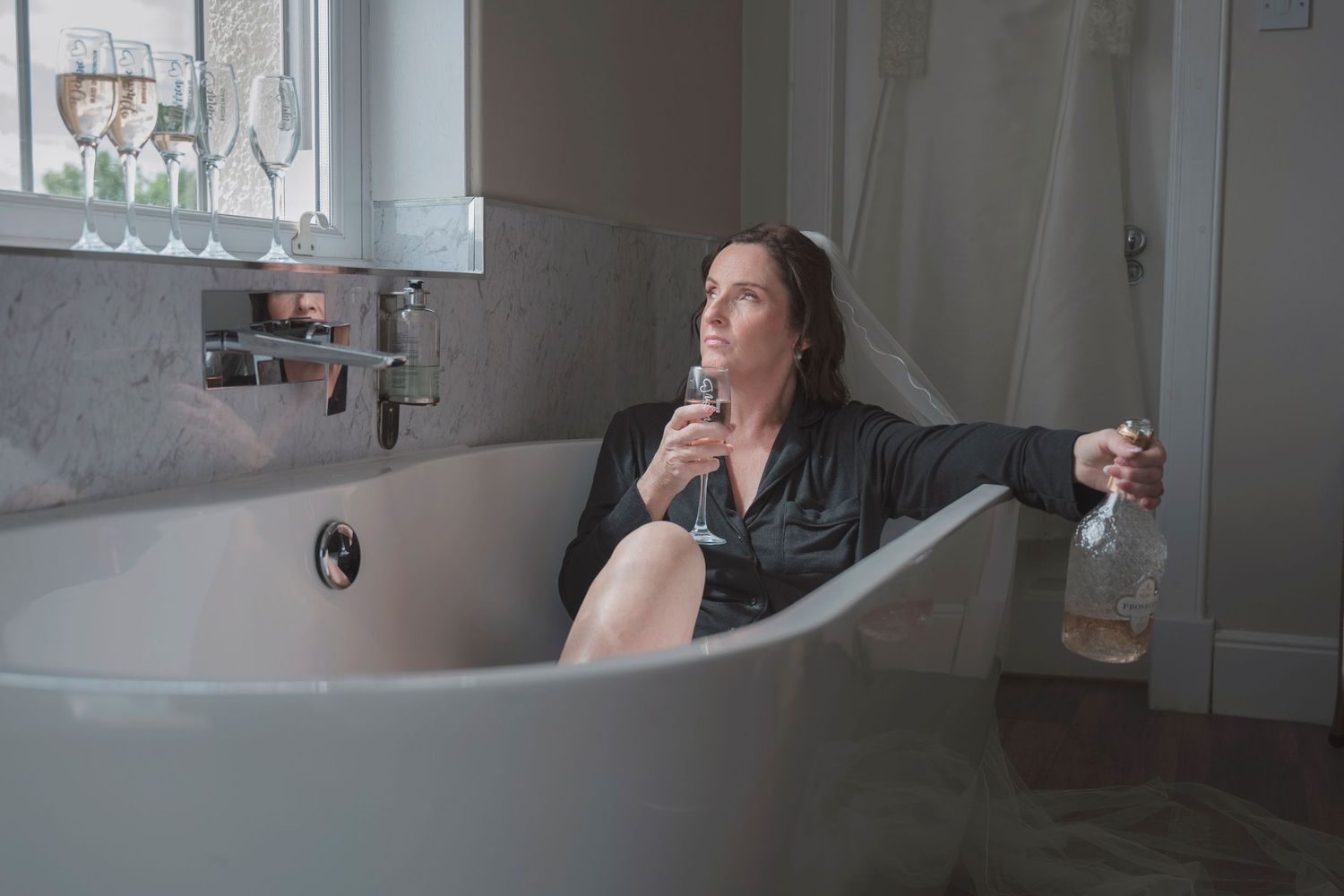Woman relaxing in freestanding bathtub holding wine glass and bottle in elegant bathroom.