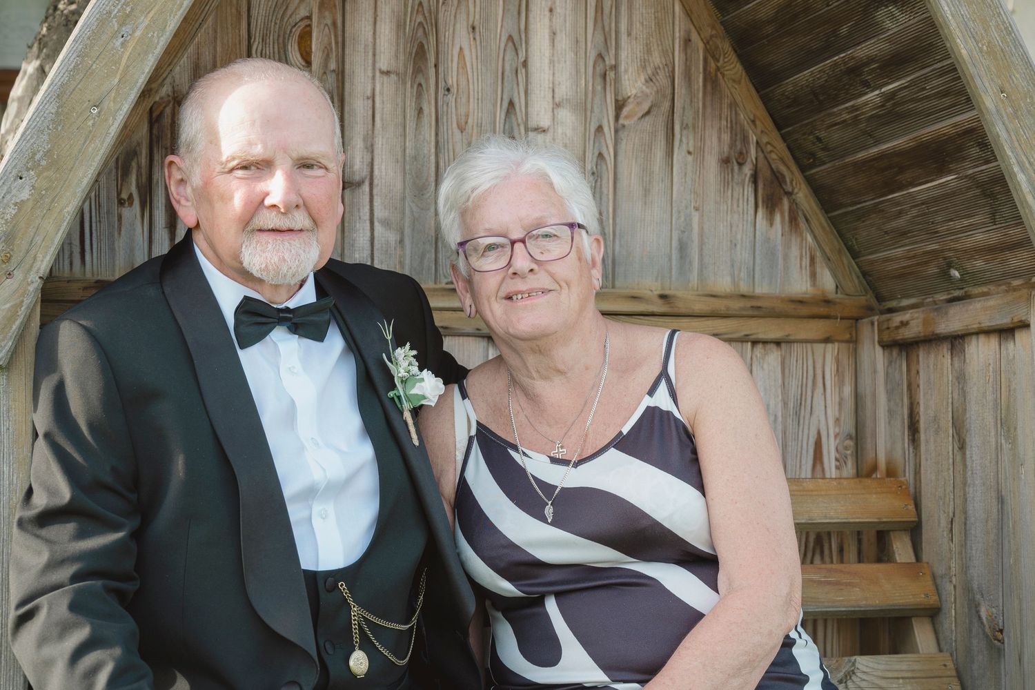 Elderly couple posing together near rustic wooden structure, man in tuxedo with boutonniere, woman in striped top.