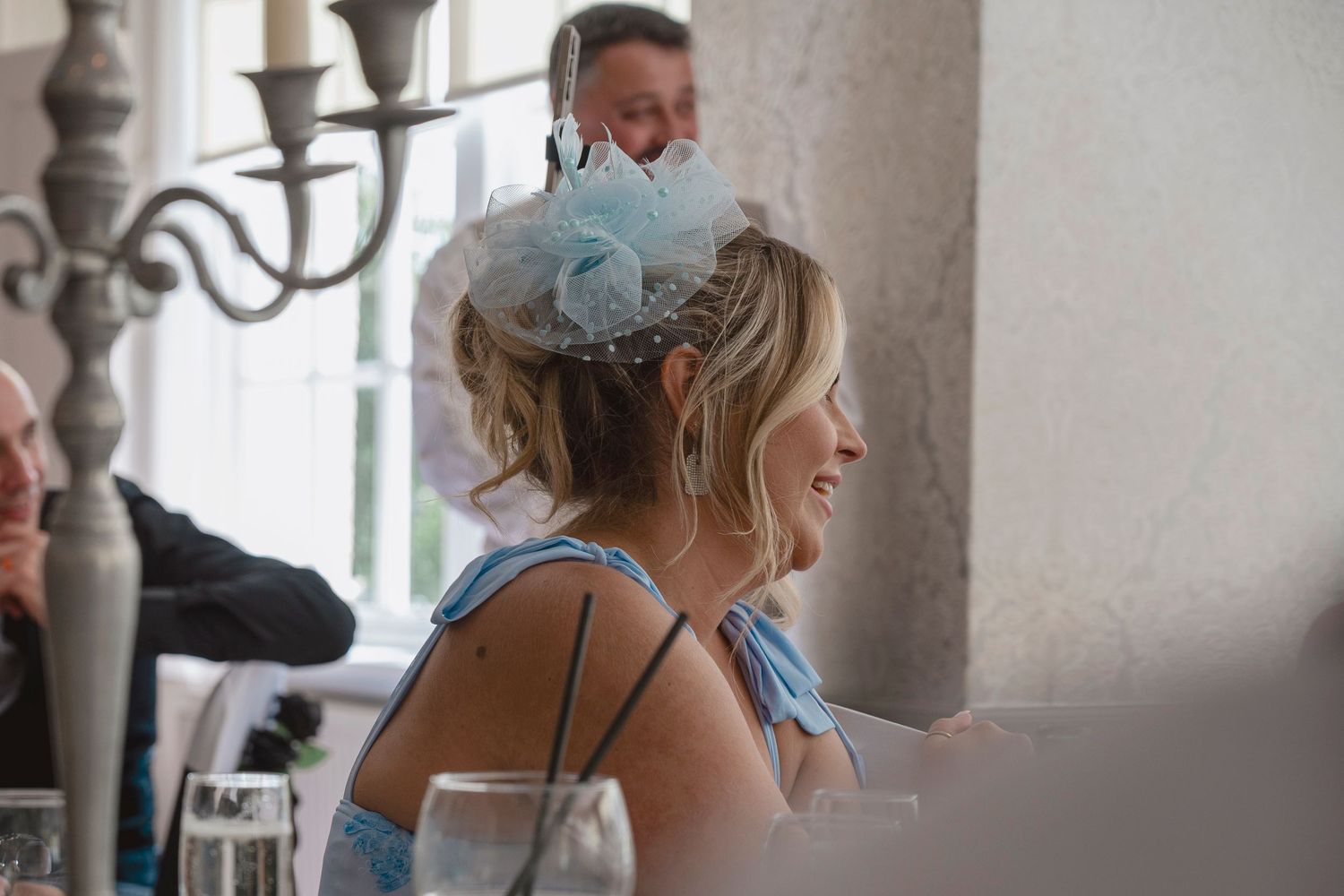 Woman wearing elegant blue fascinator hat laughing at a wedding reception dinner table.