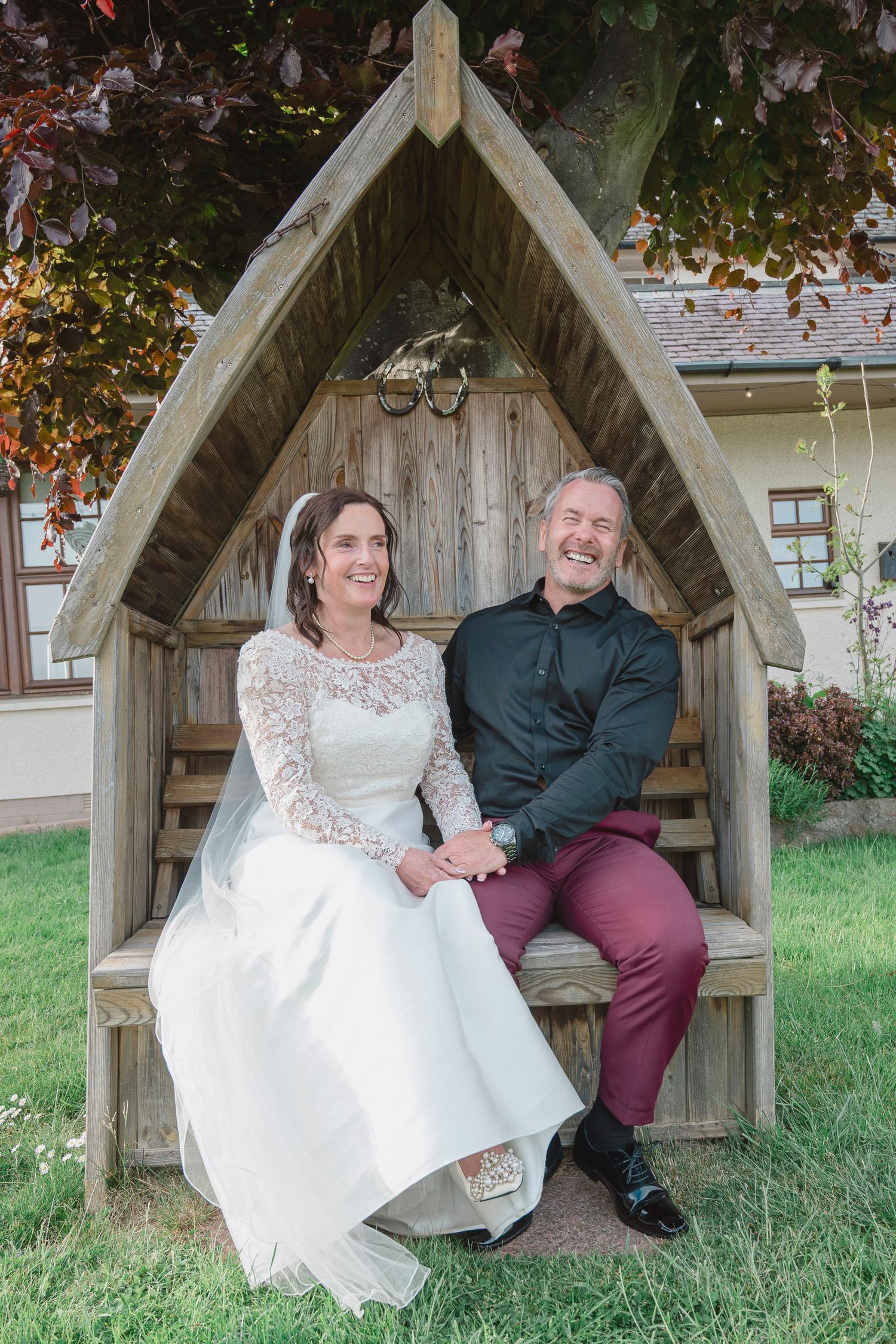 A bride and groom sit laughing together in a wooden garden arbor, she in a lace gown, he in dark shirt and burgundy trousers.
