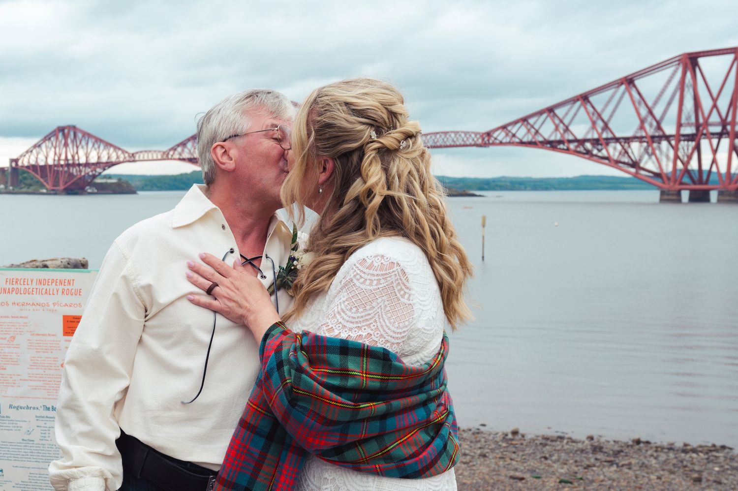 Couple kissing near the Forth Bridge in Scotland, woman in tartan shawl embracing older man by the water.