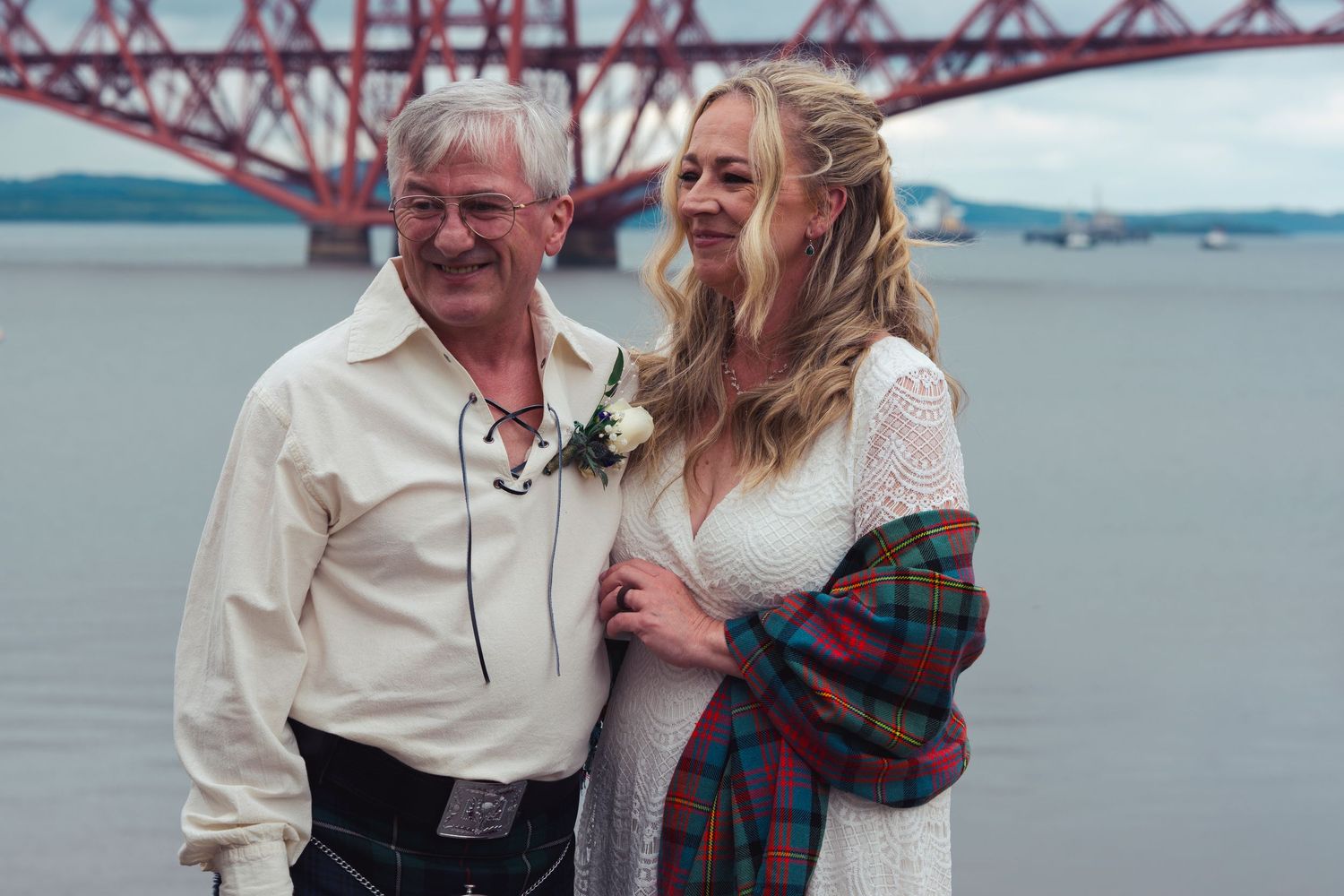 Couple in wedding attire smiling by the Forth Bridge in Scotland, bride in white dress with tartan shawl.