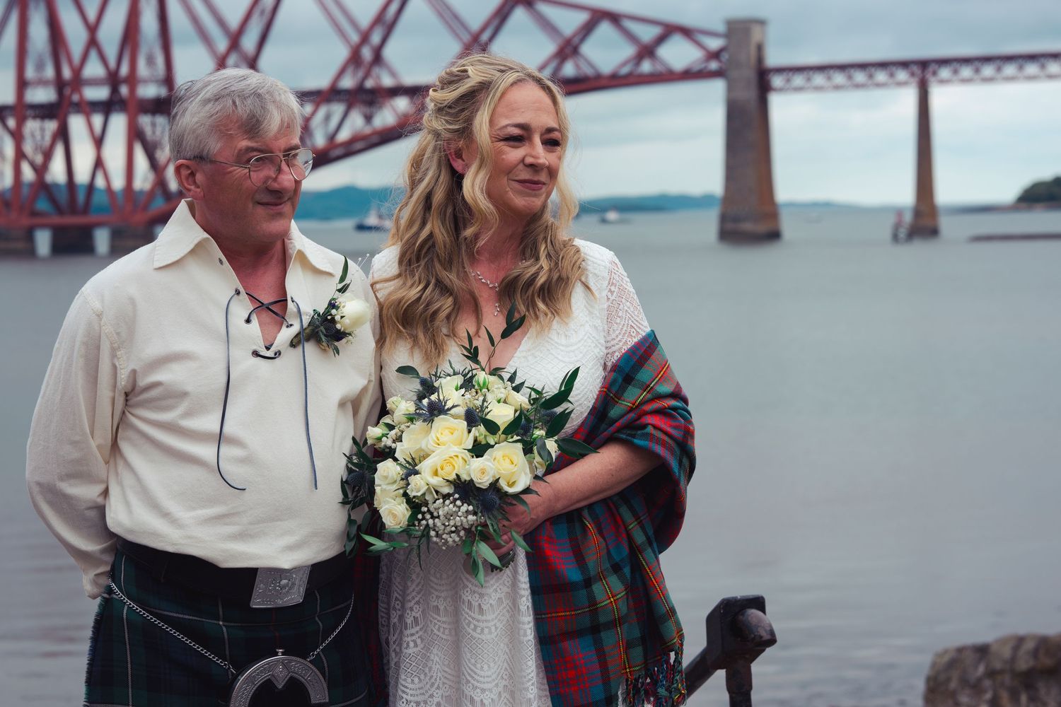 Bride holding white bouquet with groom in kilt at waterfront wedding near Forth Bridge, Scotland.
