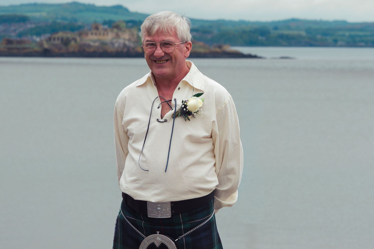 Man in Scottish kilt and cream shirt with boutonniere poses by seaside with coastal town in background.