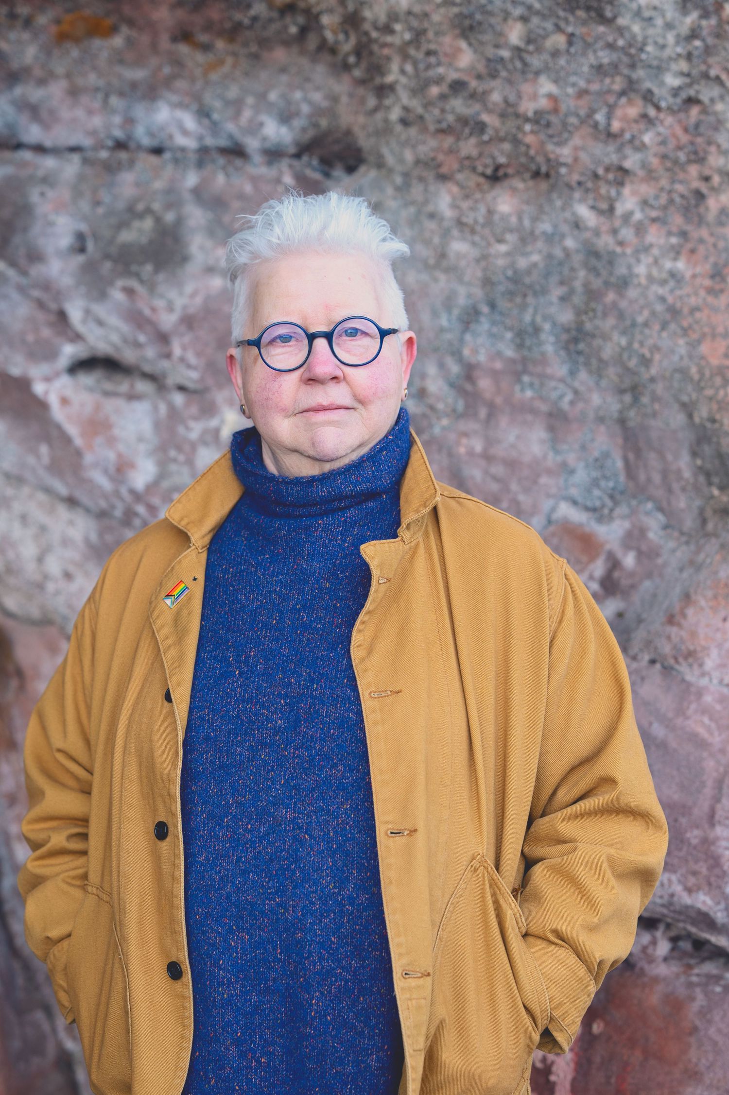 The Author Val Mcdermid with short white hair and round glasses wearing a mustard jacket and blue turtleneck sweater in the Weymss caves
