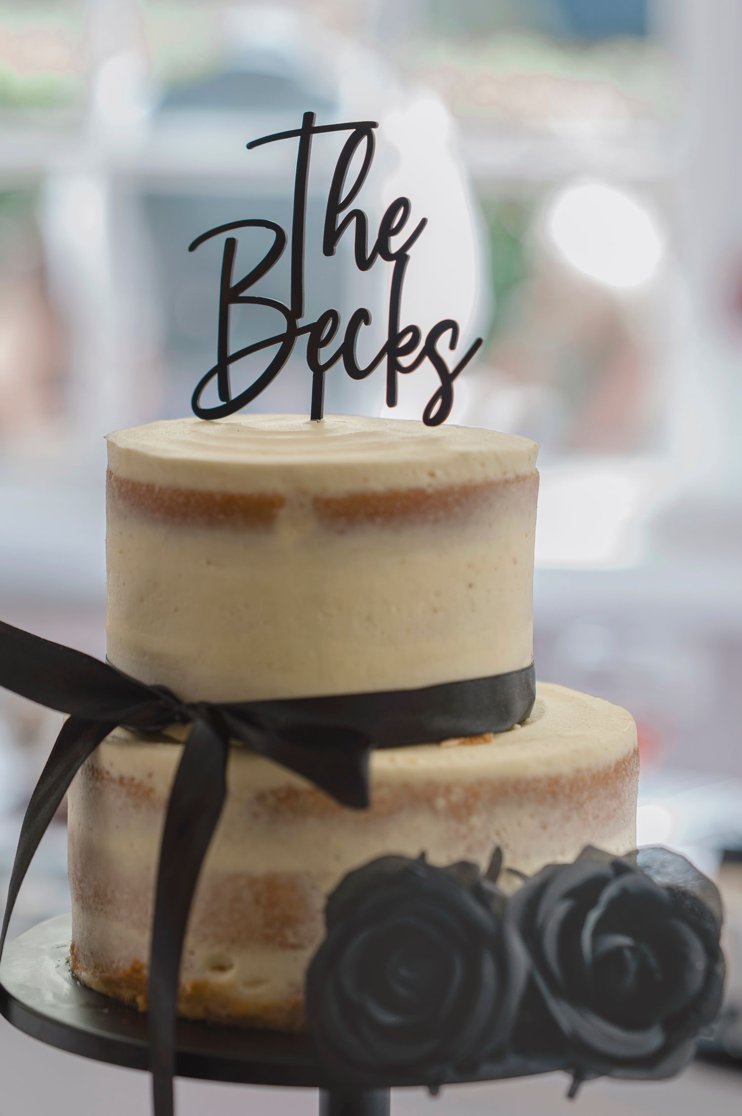 Two-tier semi-naked wedding cake with black ribbon, dark roses, and The Becks cake topper.